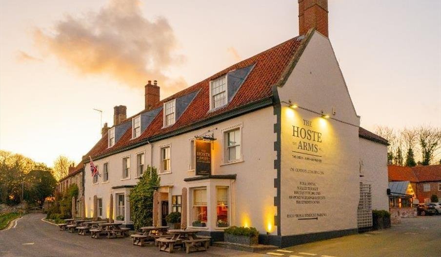 Exterior of The Hoste Arms, a white pub with red tile roof, outdoor seating with picnic tables, and illuminated sign, during sunset.
