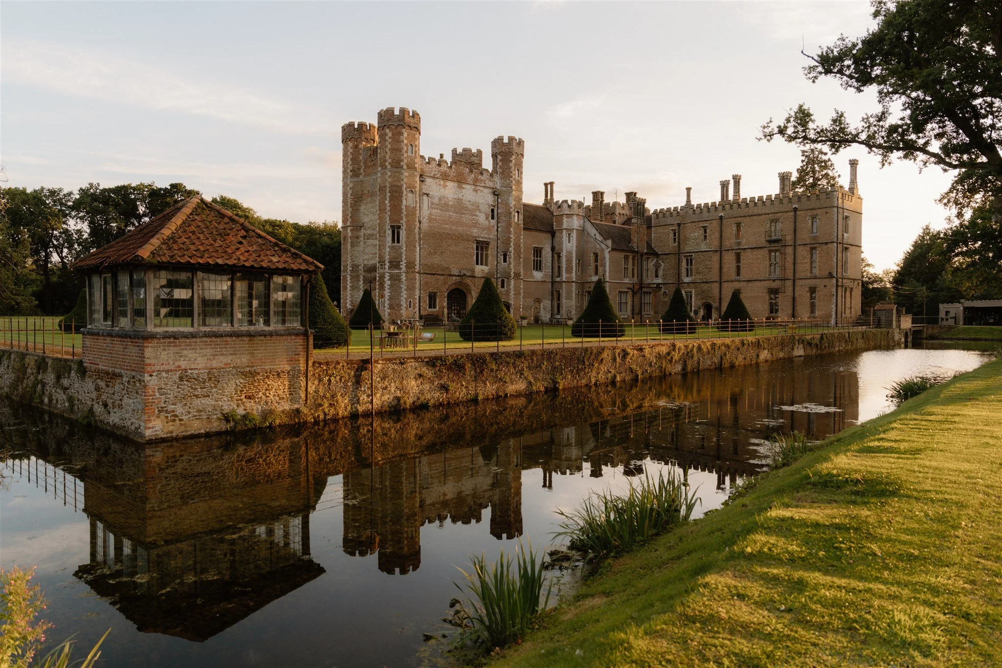 A large historic castle with multiple towers and battlements, surrounded by a moat with calm water reflecting the structure, and a small brick building with a tiled roof in the foreground, set against a backdrop of green trees and a partly cloudy sky.