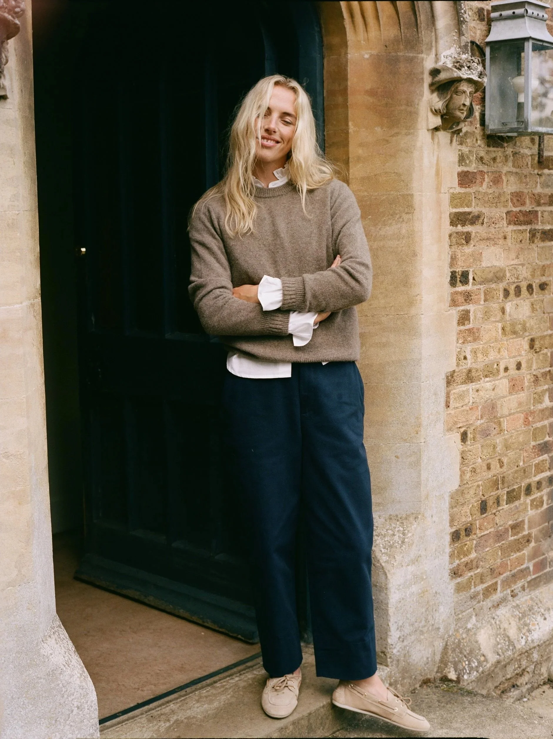 A woman with long blonde hair wearing a grey sweater, white shirt, and navy pants standing at a doorway in front of an old brick wall with a stone face sculpture and a glass lantern.