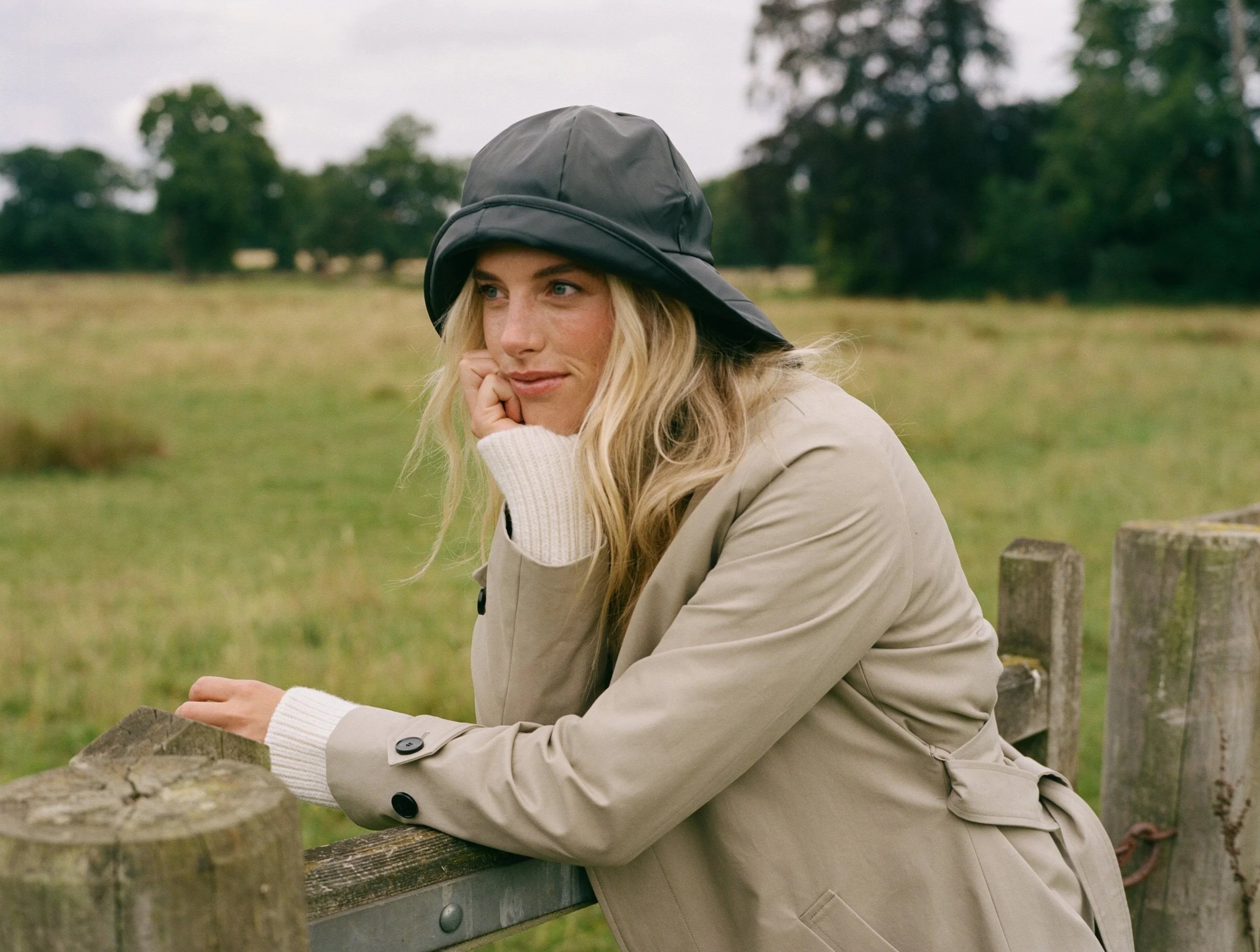 A woman with blonde hair wearing a black rain hat and beige trench coat sitting on a wooden fence in a grassy field with trees in the background.