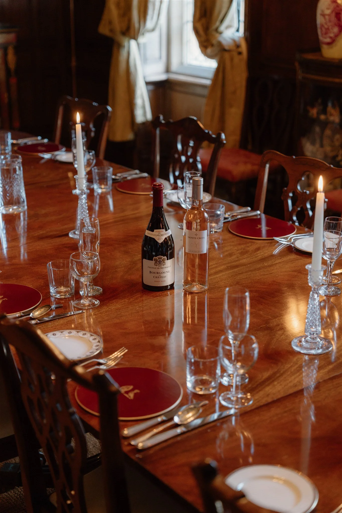 A dining room table set for a meal with red plates, silverware, wine bottles, lit candles, and glasses, with wooden chairs around and a window with curtains in the background.