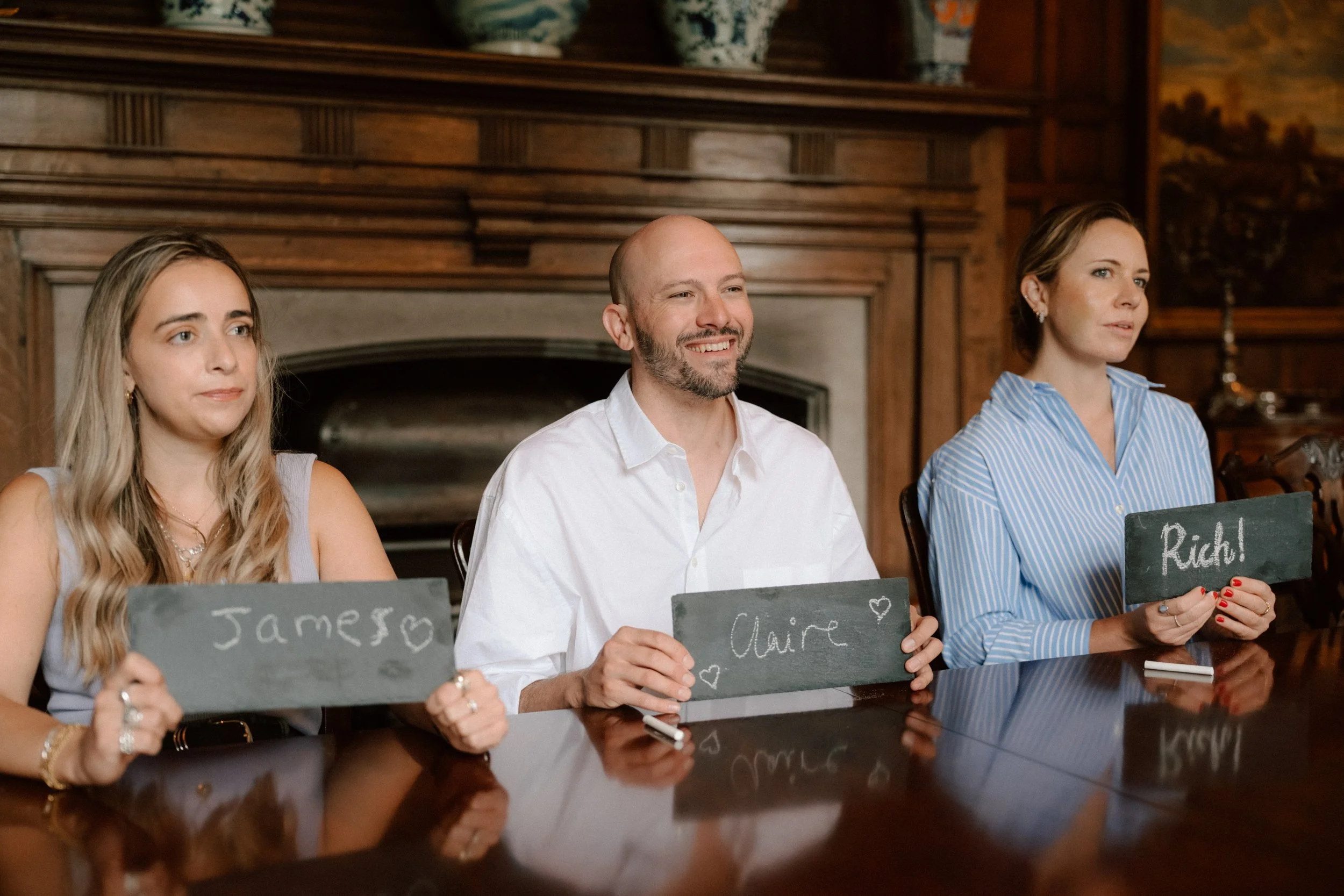Three people sitting at a table, each holding a small chalkboard sign with handwritten names and hearts. The woman on the left has long blonde hair, the man in the middle has a bald head and beard, and the woman on the right has brown hair tied back. They are in a wood-paneled room with a fireplace and decorative pottery on the mantle.