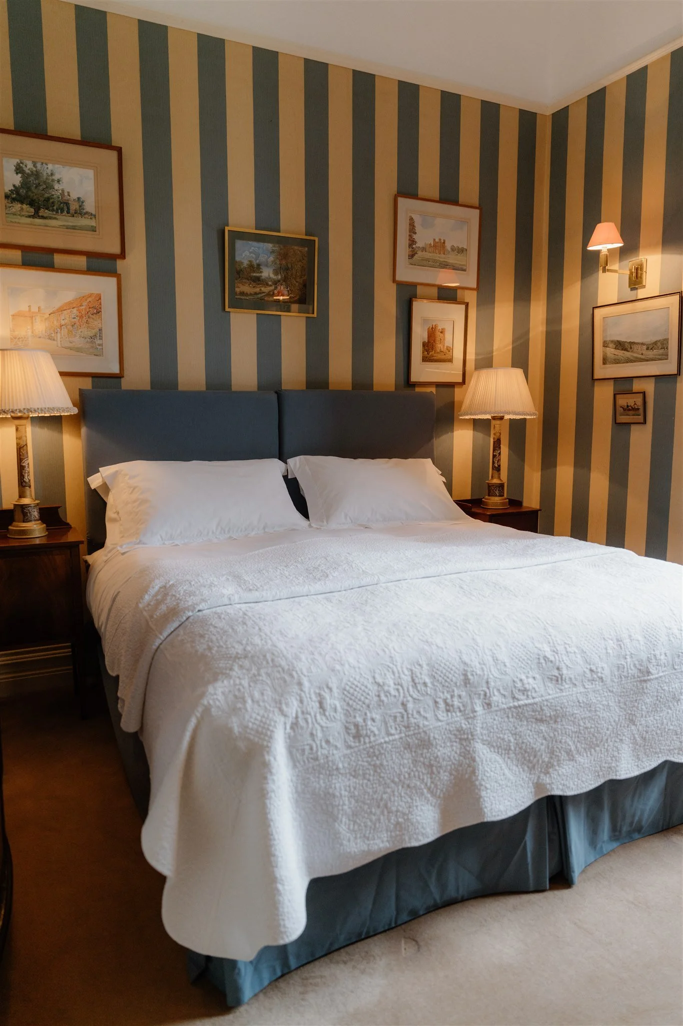 A neatly made bed with white quilted bedspread, two pillows, blue upholstered headboard, and wooden bedside tables with table lamps, situated in a room with striped yellow and blue wallpaper and framed landscape paintings.