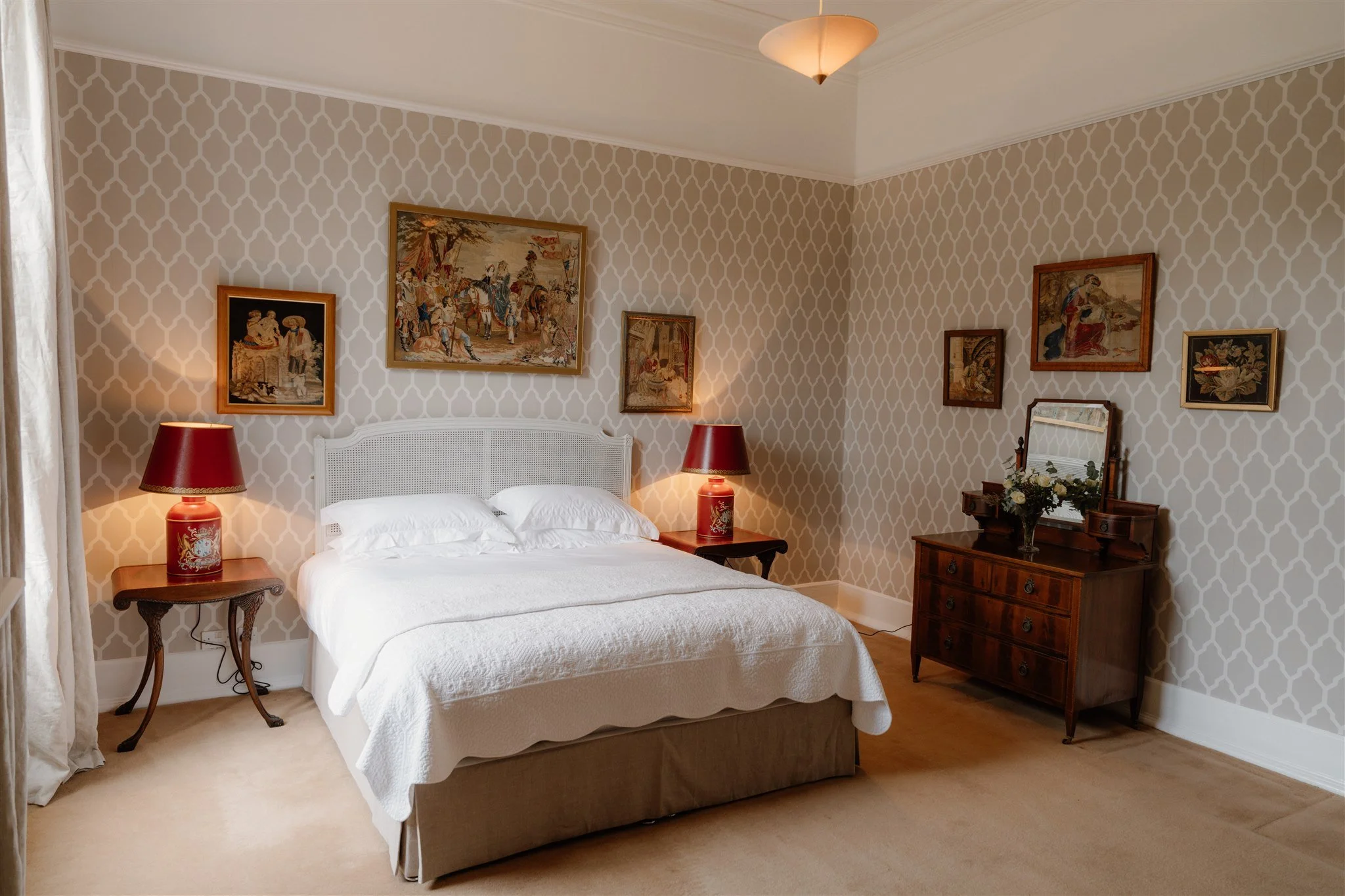 A cozy bedroom with a white bed, red lamps on wooden nightstands, framed artwork on patterned wallpapered walls, and a wooden dresser with a mirror and flower arrangement.