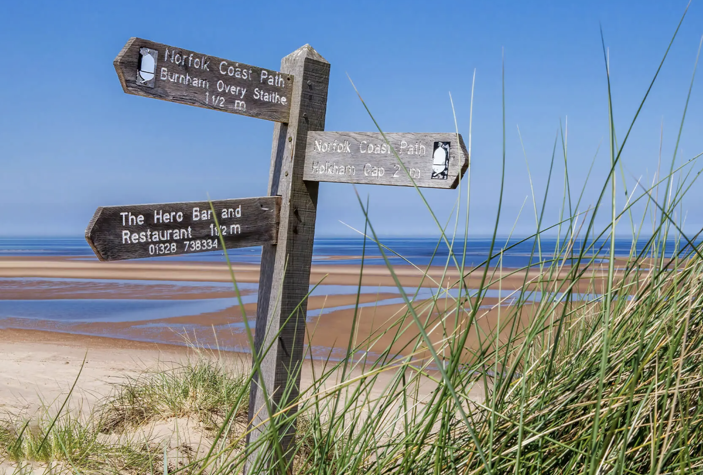 Wooden signs marked with trail information and distances near a beach with sand, water, and tall grass.