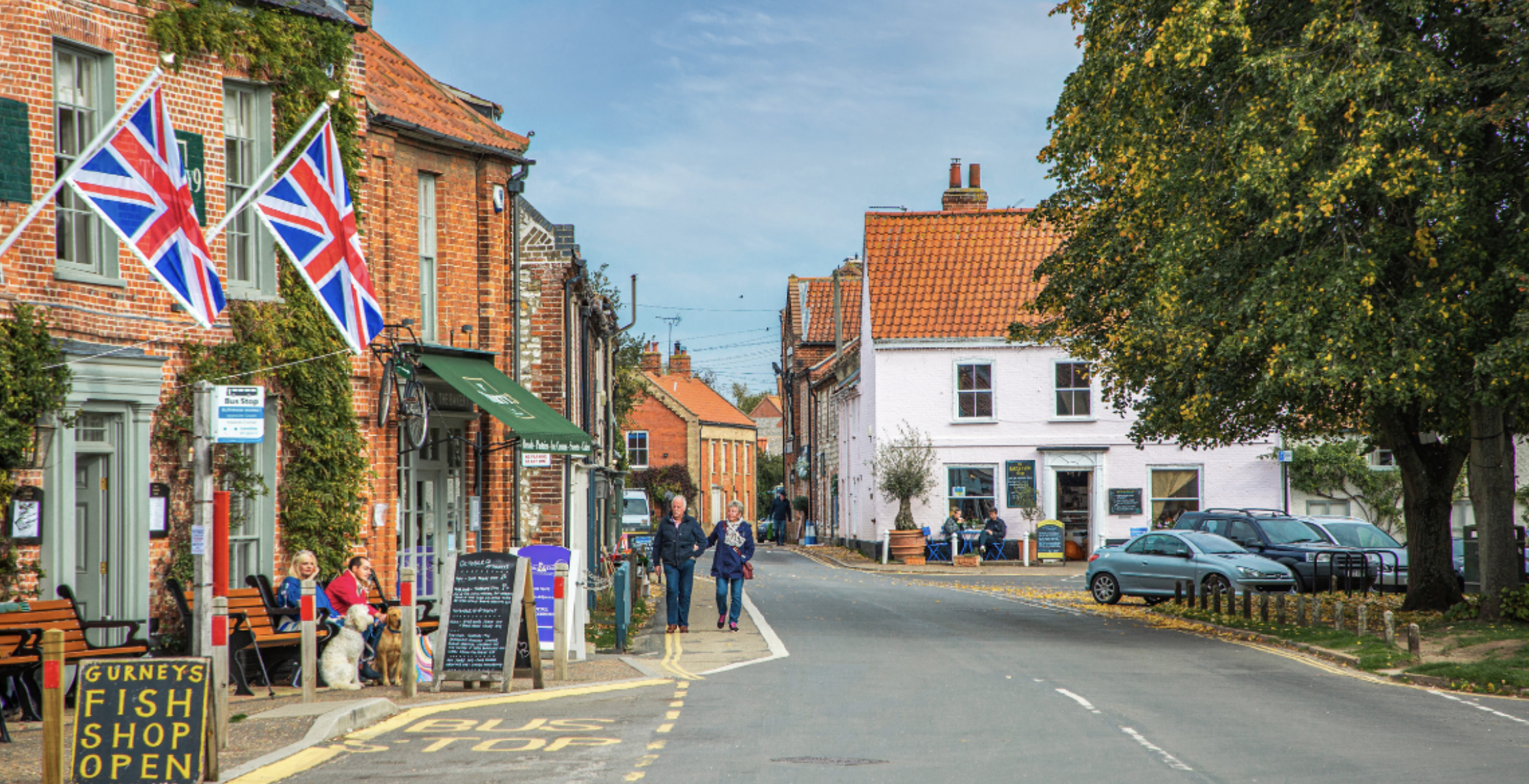 A charming street scene in a small town with outdoor seating, shops, and people walking. The buildings are brick with some greenery, shops have signs and flags, and cars are parked along the street. The sky is partly cloudy with some trees displaying fall foliage.