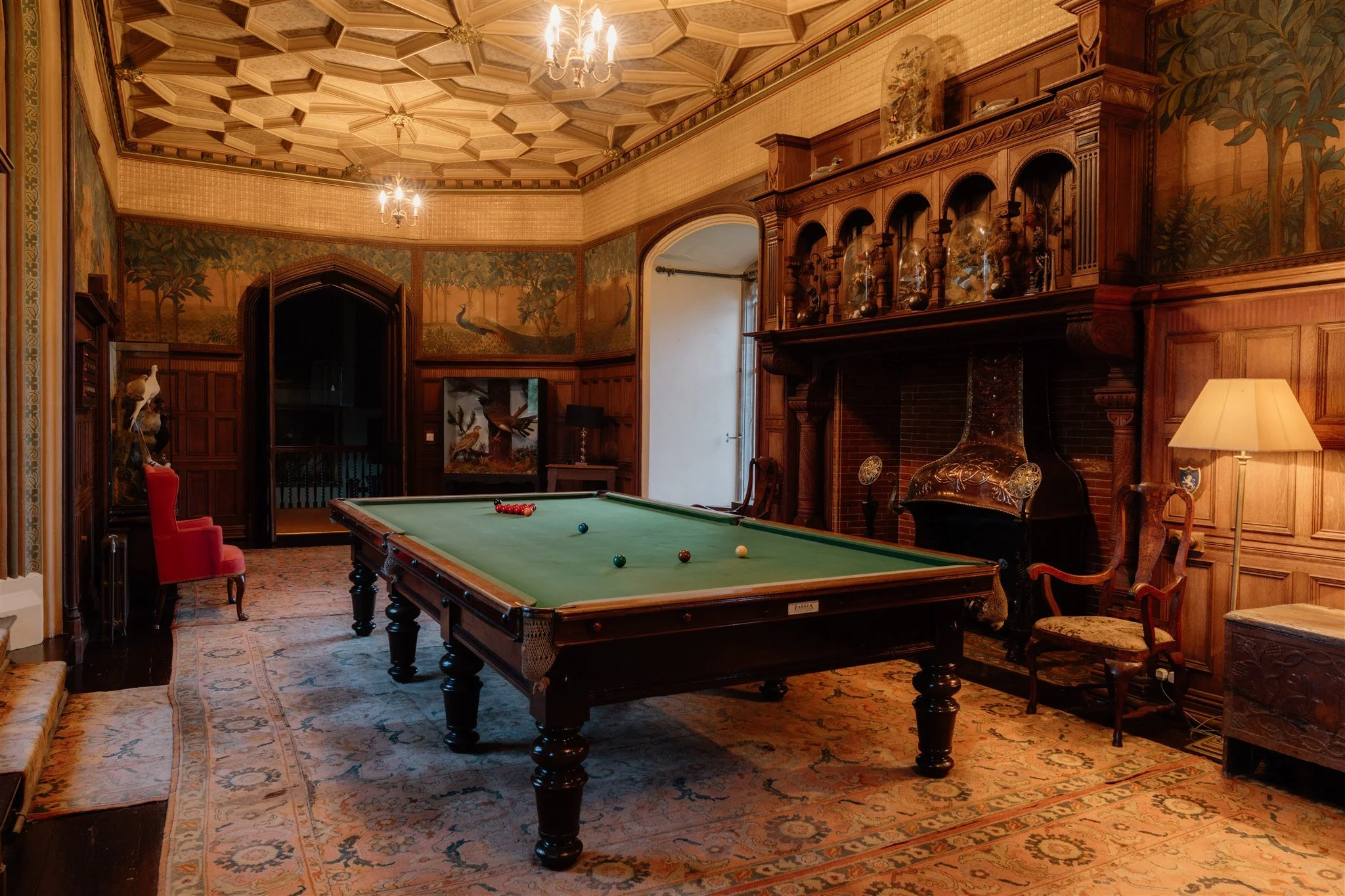 A vintage-style room with a green billiards table, ornate wood paneling, a large fireplace, wall paintings of peacocks and trees, chandeliers, and a patterned carpet.
