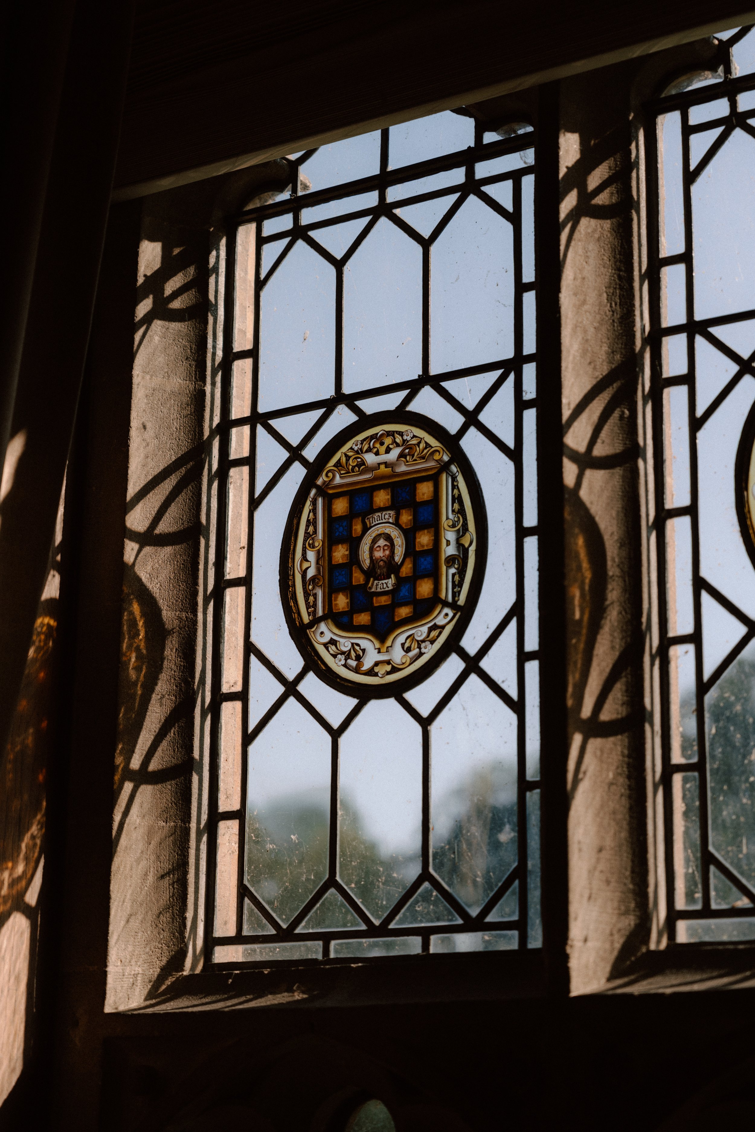 View through a decorative stained glass window showing a coat of arms with a saint's face on a blue background, casting shadows inside a stone building.