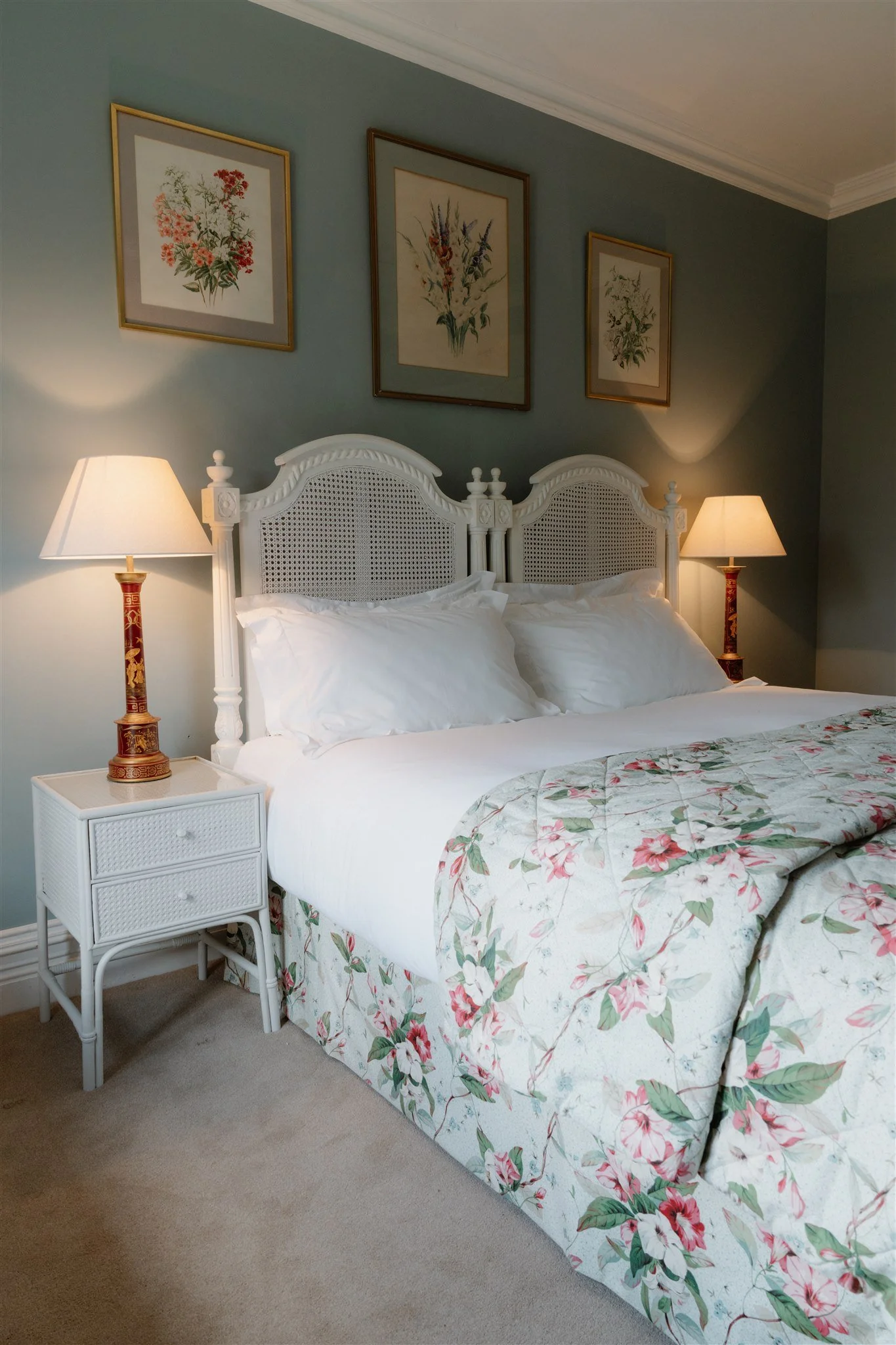 A bedroom with a white bed featuring a floral bedspread, two pillows, and a carved headboard. Two bedside tables with lamps are on each side of the bed. Above the bed, three framed floral art prints hang on a blue wall.