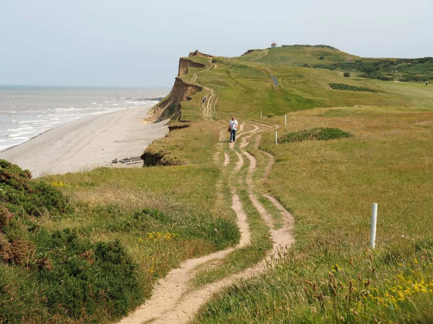 A dirt path along grassy terrain leading to a cliffside overlooking a beach and ocean, with a person walking along the trail and a lighthouse at the top of the hill.