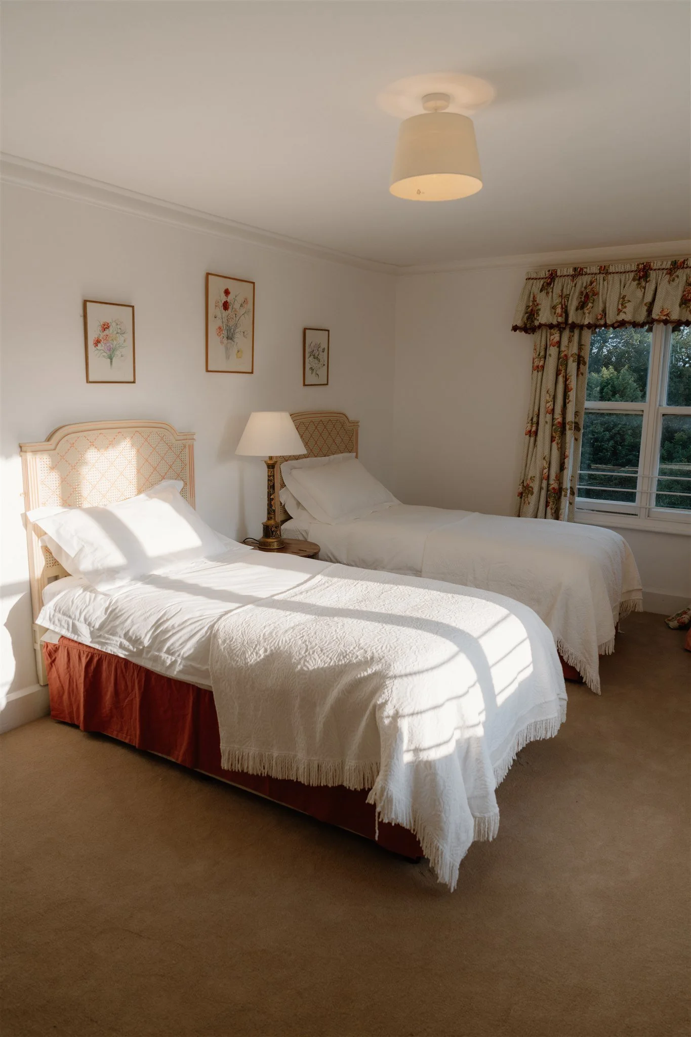 A hotel room with two single beds, each with white bedding and a pillow. There is a nightstand between the beds with a lamp. The wall behind the beds has three framed floral artworks. A window with floral curtains shows trees outside. The room has beige carpet and a ceiling light fixture.