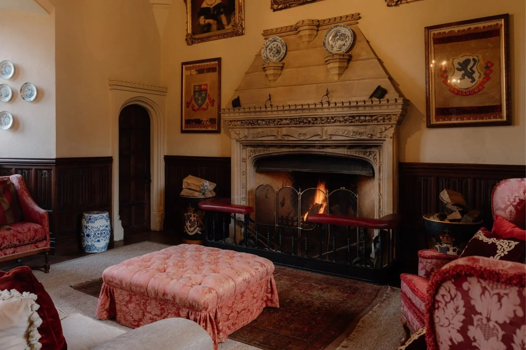 A cozy living room with a lit fireplace, vintage pink and red upholstered furniture, framed historical illustrations on the wall, and decorative plates. There's an ottoman and a small table near the fireplace, with logs stored beside it.