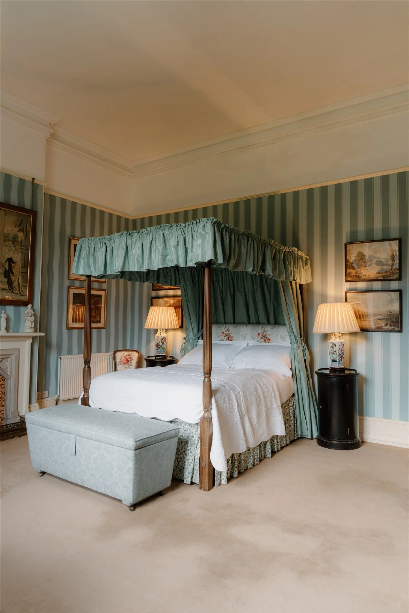 A bedroom with a four-poster bed draped with green curtains and a floral headboard, flanked by two matching bedside lamps. There is a small upholstered ottoman at the foot of the bed, and the room features framed landscape and portrait paintings on striped wallpapered walls, with a white fireplace and decorative figurines on the mantel.