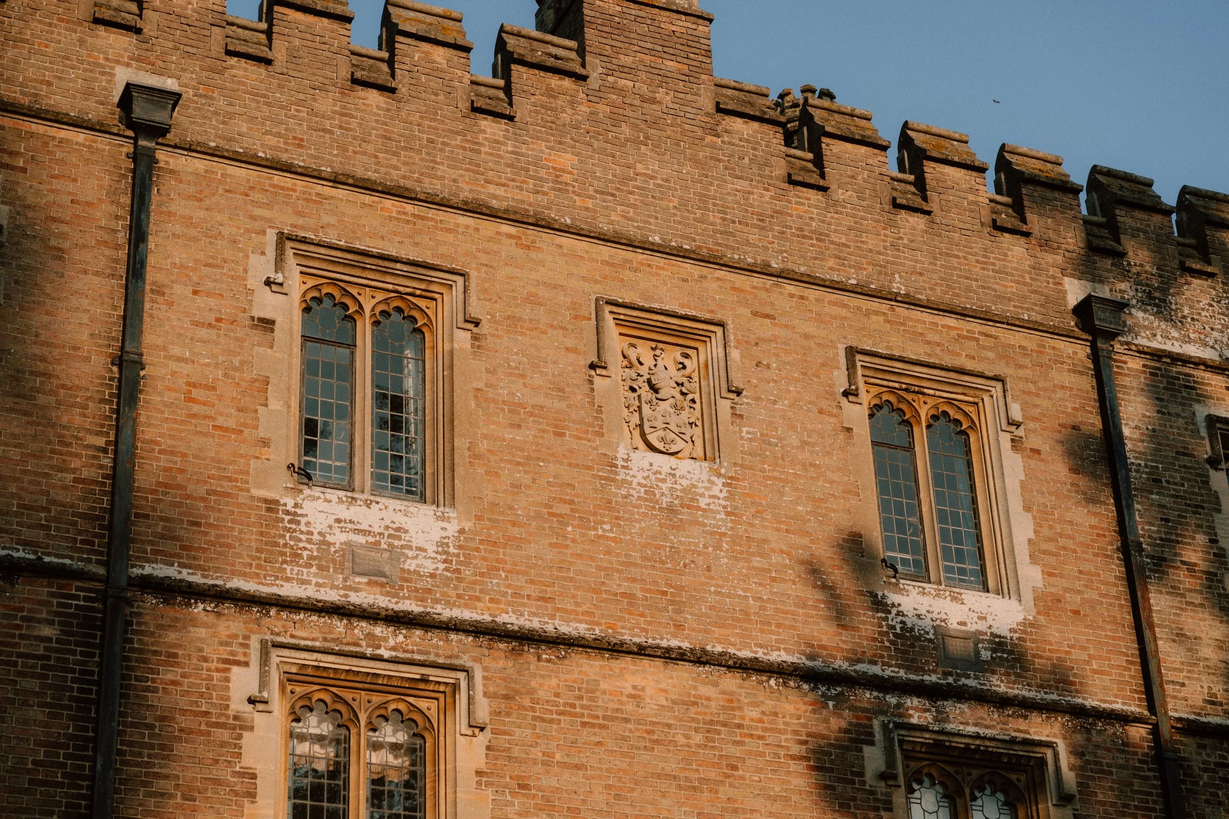 A close-up view of a historic brick building with ornate window frames and a coat of arms carved in stone on the wall. The building has gothic-style windows with decorative tracery, and a weathered appearance with some peeling paint and moss on the bricks. The sky is clear and blue.