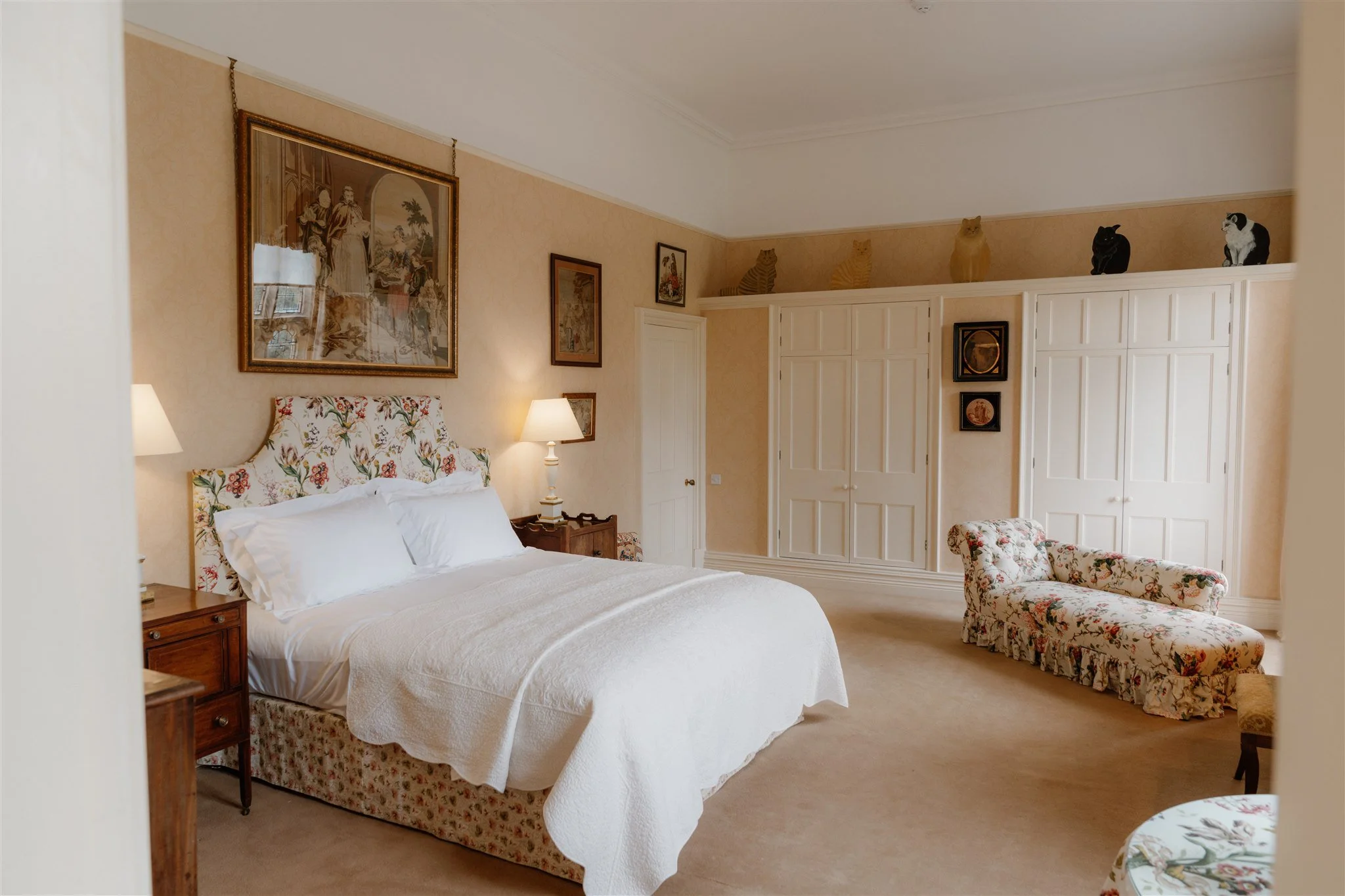 A bedroom with a bed featuring a floral headboard, a nightstand with a lamp, framed artwork on the wall, a white closet with wooden doors, a floral armchair, and animal figurines of cats and dogs on top of the closet.