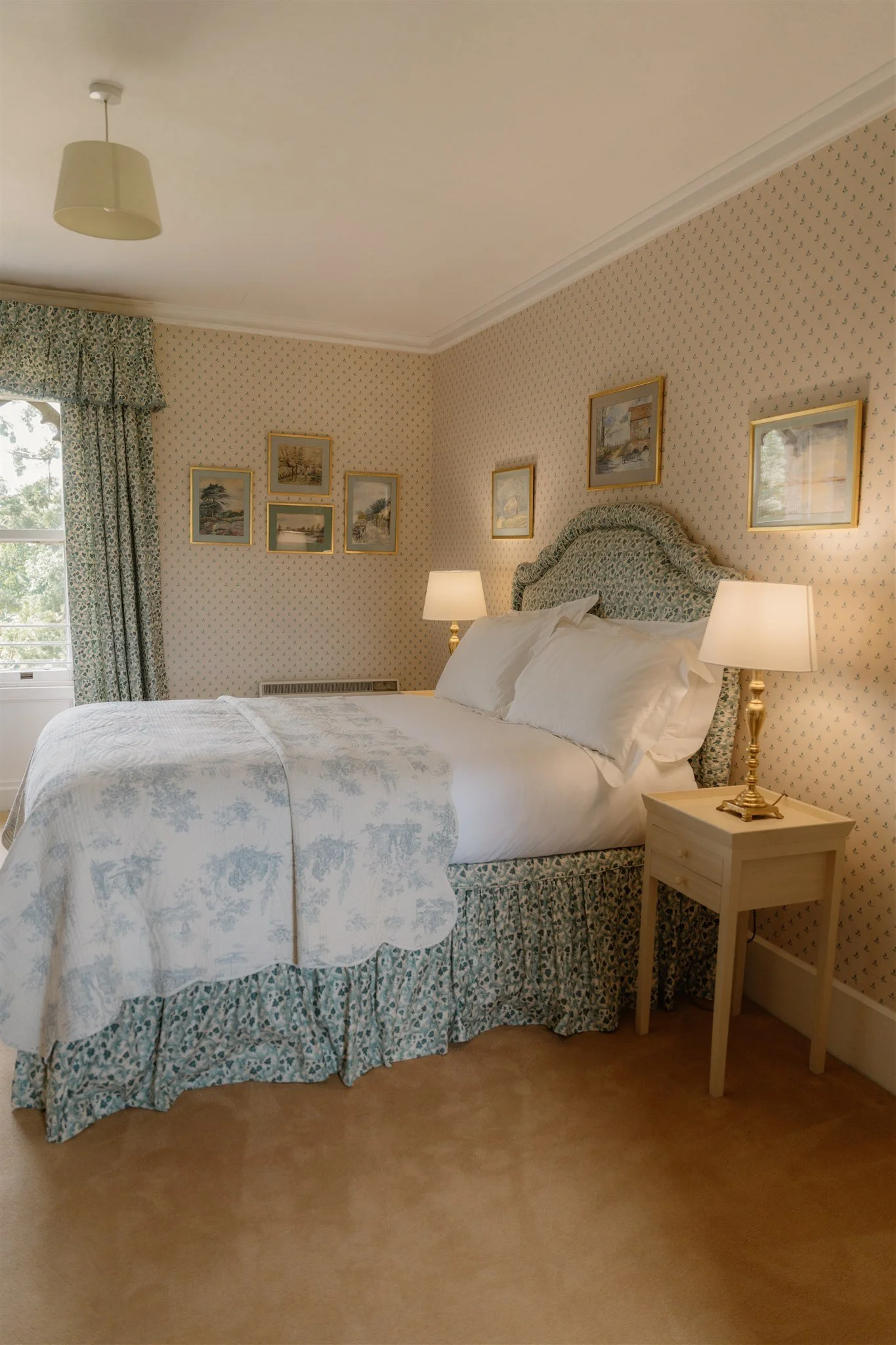 A cozy bedroom with a large bed that has a floral-patterned headboard, white bedding, and matching curtains. There are two bedside lamps with gold bases on light-colored wooden nightstands, and framed artwork hanging on the wall above the bed. The room has beige carpeting and light-colored walls with small, patterned wallpaper.