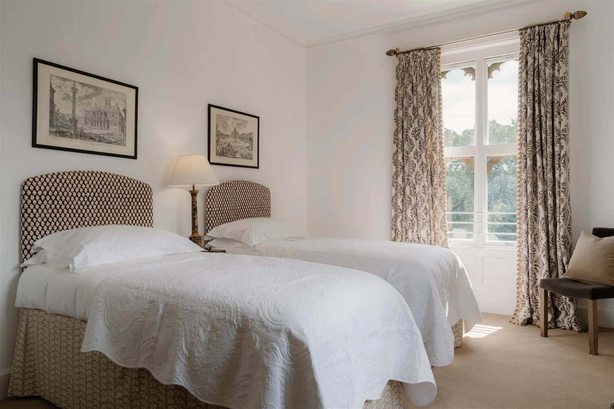 A bedroom with two beds, white bedding, patterned headboards, a table lamp between the beds, and a large window with patterned curtains, overlooking trees outside.