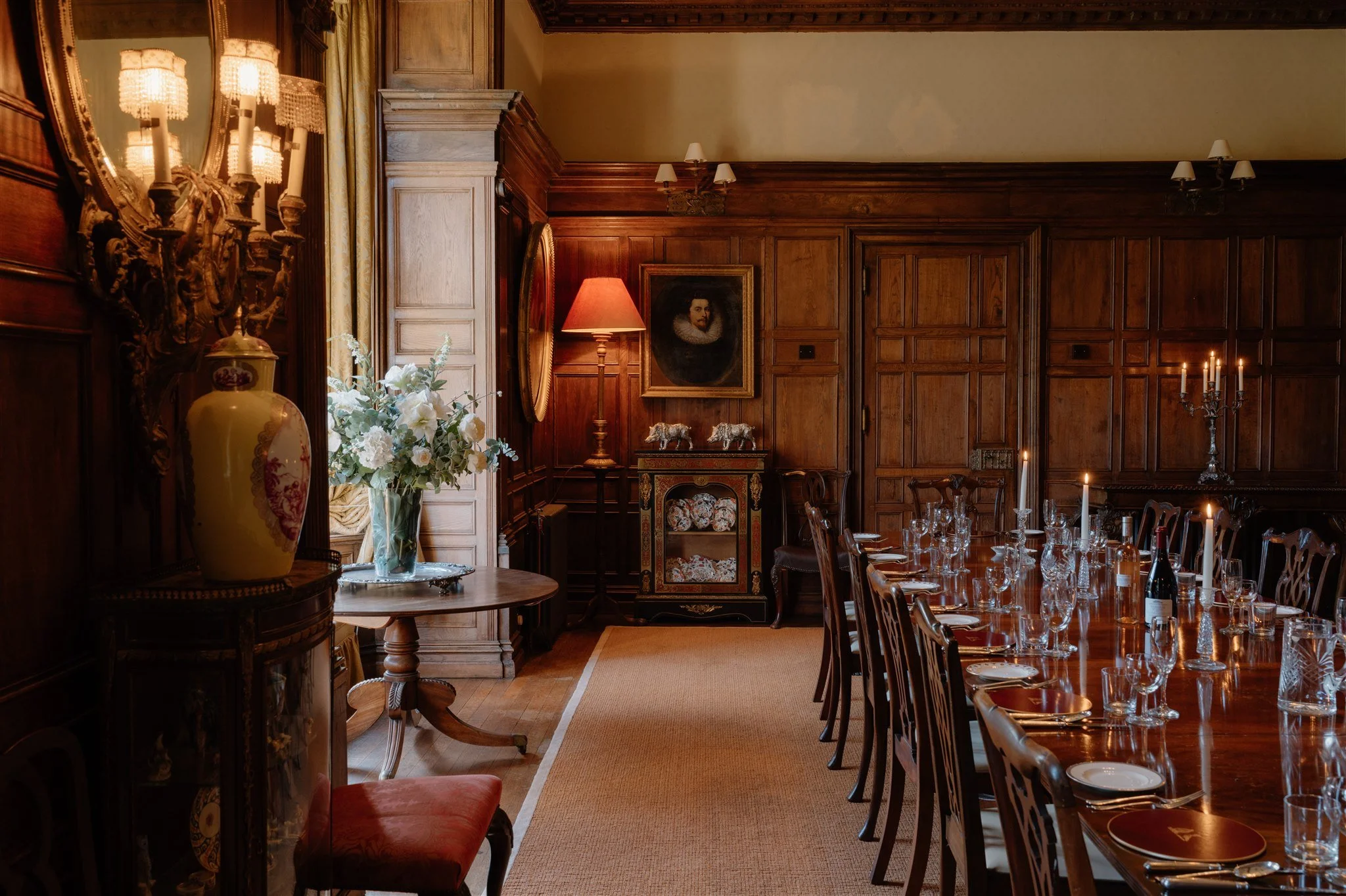 A formal dining room with wood-paneled walls, a large wooden dining table set with glassware and candles, ornate wall sconces, a portrait of a man with a ruffled collar, a bouquet of white flowers on a small table, and classic furnishings with a warm, elegant atmosphere.