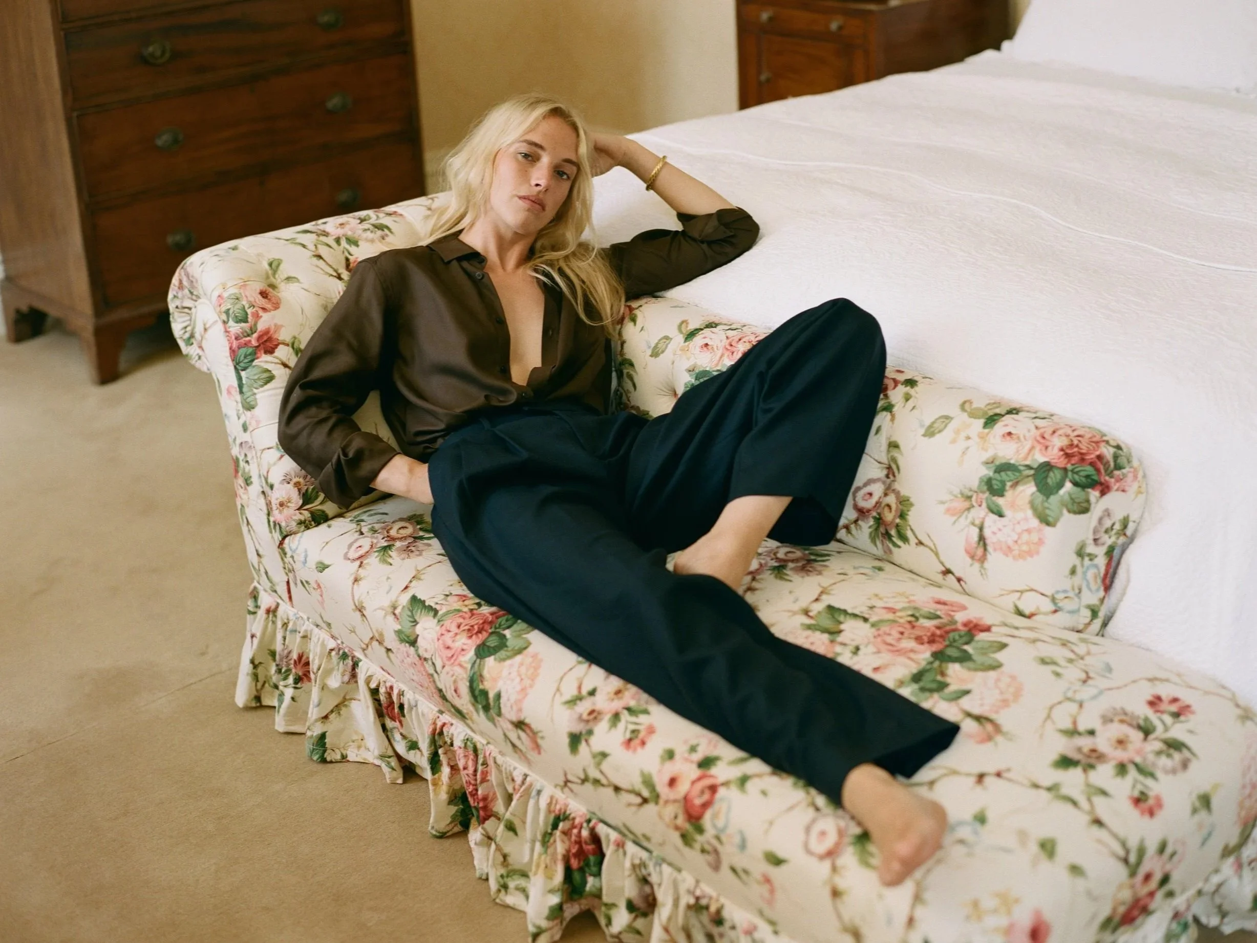 A woman with blonde hair, wearing a dark silk blouse and black pants, sitting on a floral upholstered sofa in a room with wooden furniture and a beige carpet.