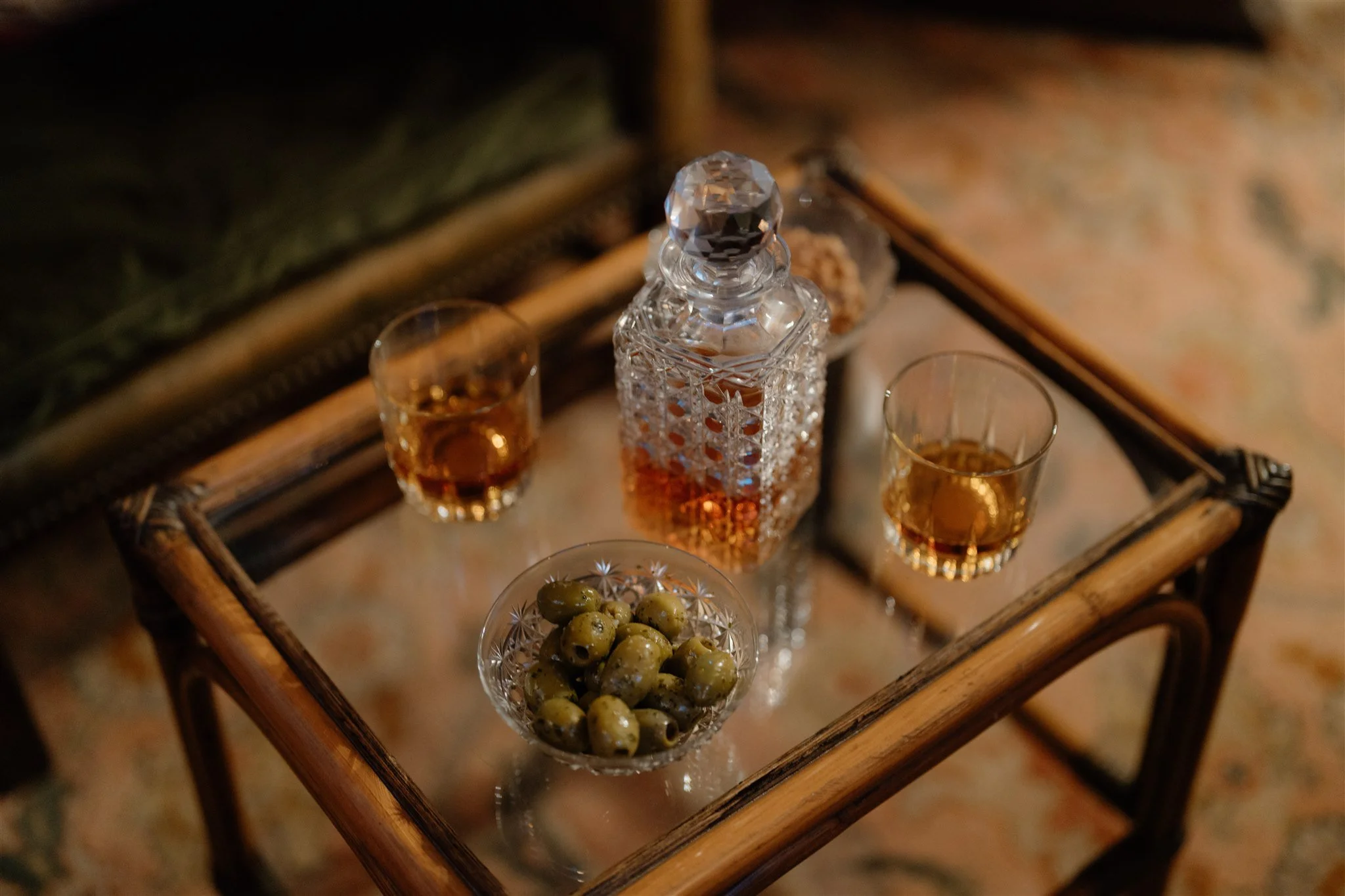 A glass decanter and two glasses with amber-colored liquid on a wicker tray, along with a small bowl of green olives, all arranged on a surface.