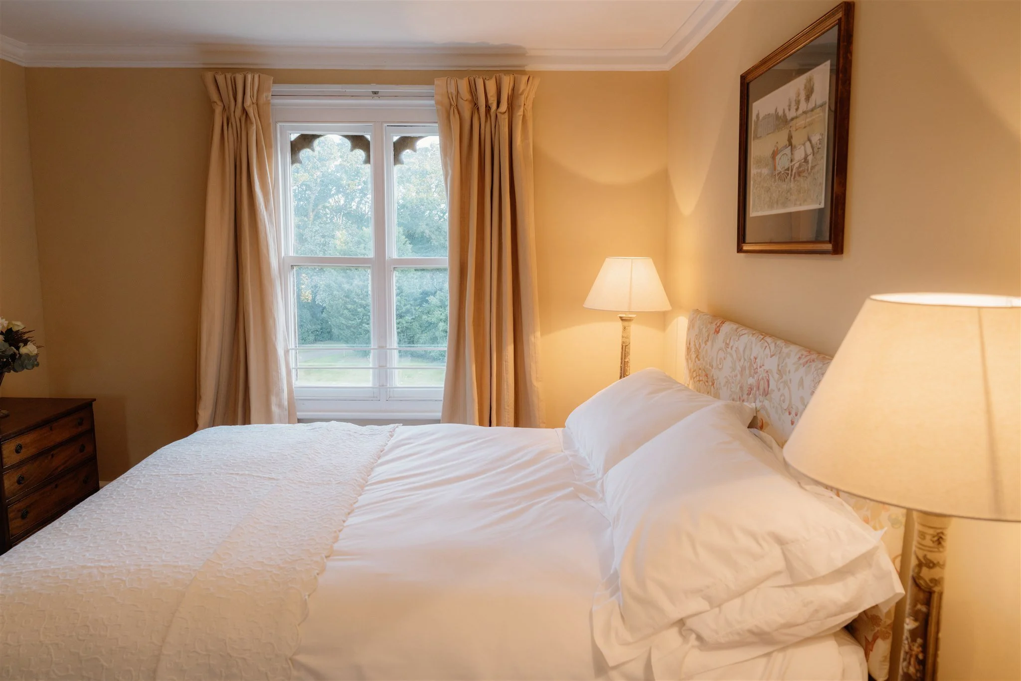A cozy bedroom with a bed, white bedding, a floral-patterned headboard, a wooden nightstand, a table lamp, a window with beige curtains, a painting on the wall, and trees visible outside.