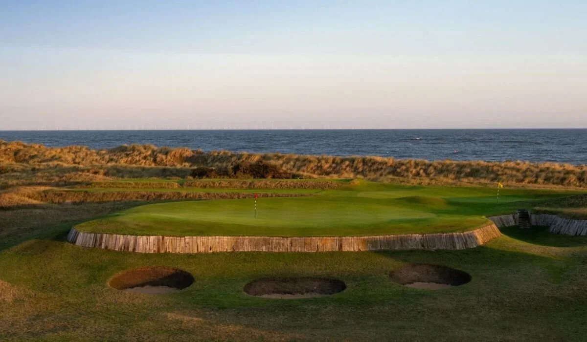 Golf course green near the ocean with sand dunes in the background and a blue sky.