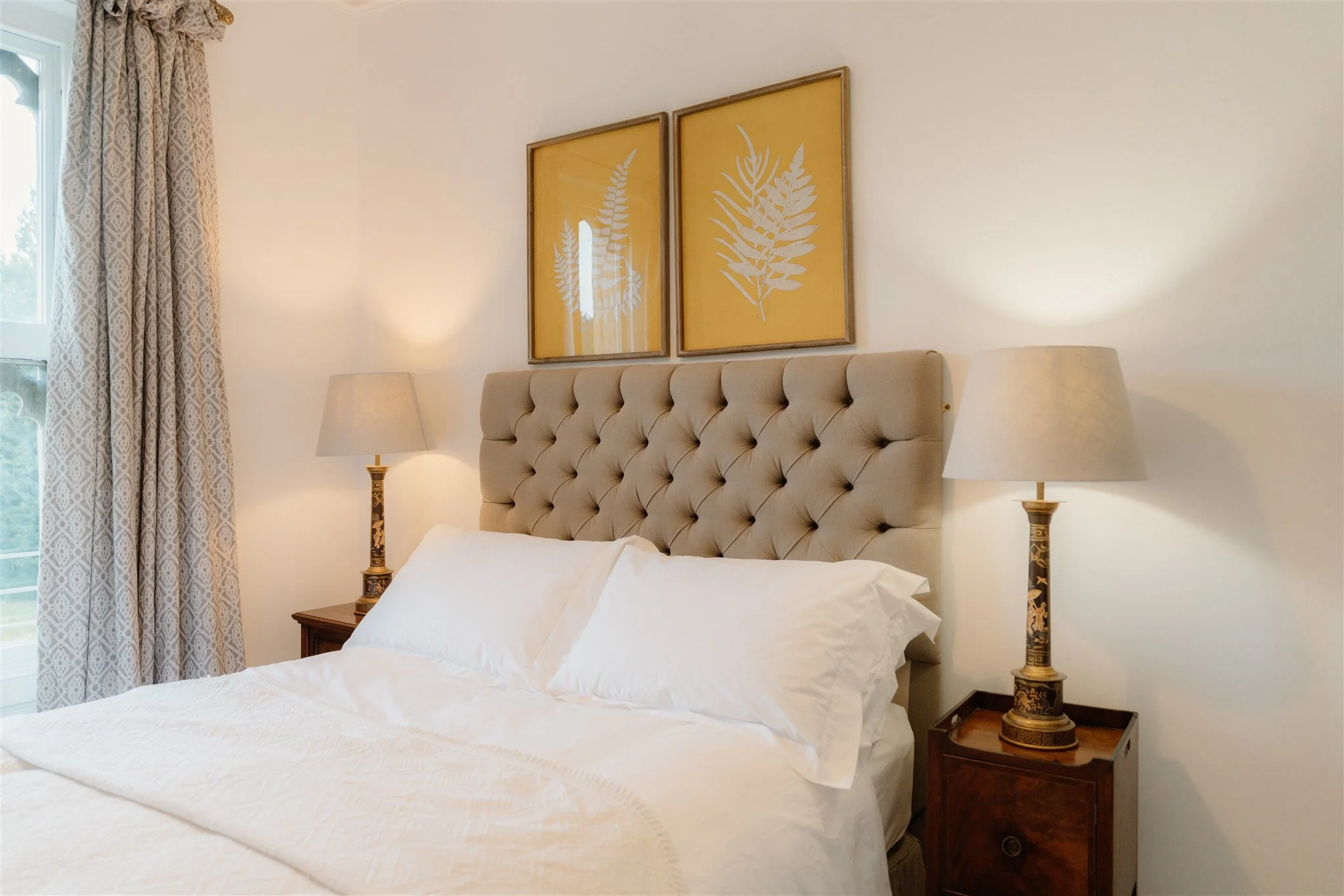 A bedroom with a beige tufted headboard, two nightstands with ornate lamps, and two framed botanical artwork prints of white fern leaves on yellow backgrounds hanging above the bed. There are beige patterned curtains beside a window on the left.