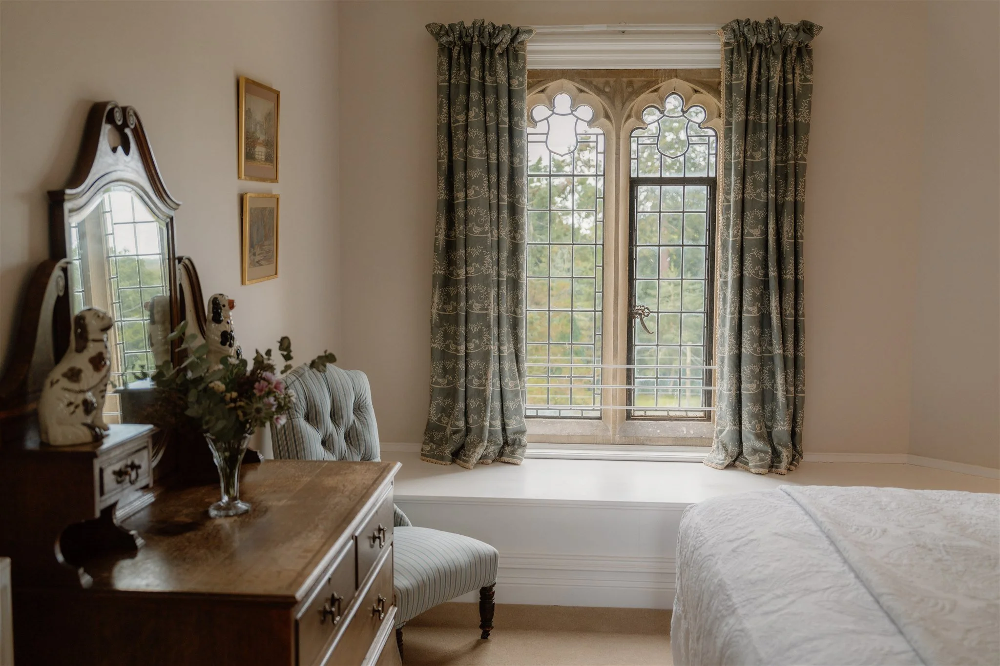 A bedroom with large stained glass windows, patterned blue curtains, a bed with a white bedspread, a wooden dresser with a mirror, and decorative porcelain dogs on the dresser.