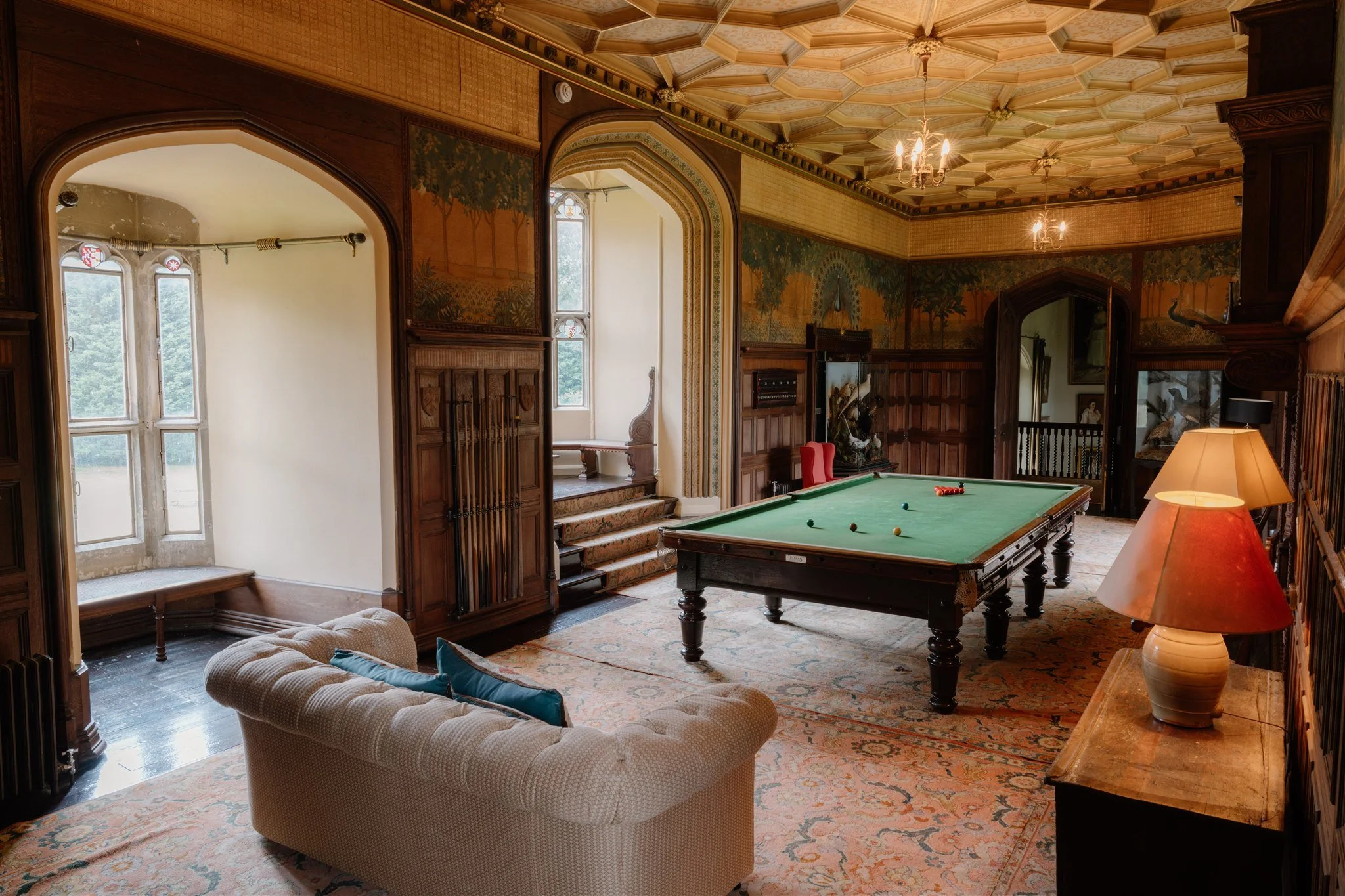 A vintage-style game room with a green pool table, a beige tufted sofa with pillows, wooden paneling, and large windows. The room has ornate ceiling detailing, chandeliers, and decorative paintings of peacocks and nature scenes on the walls.