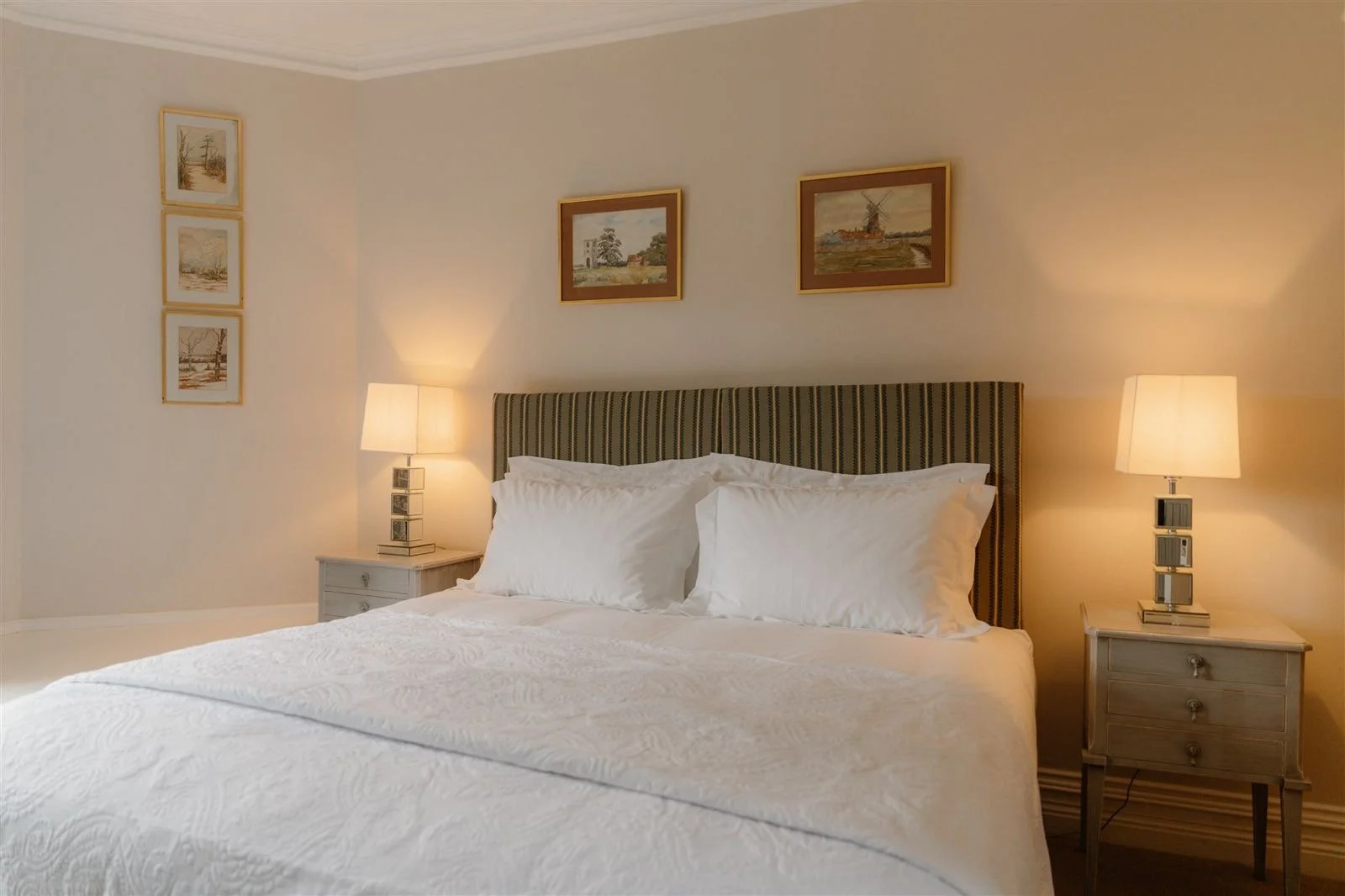A neatly made bed with white linens and pillows in a bedroom. Two nightstands with lamps are on each side of the bed, and framed artwork hangs above the headboard. The wall is painted light cream, creating a cozy and inviting atmosphere.
