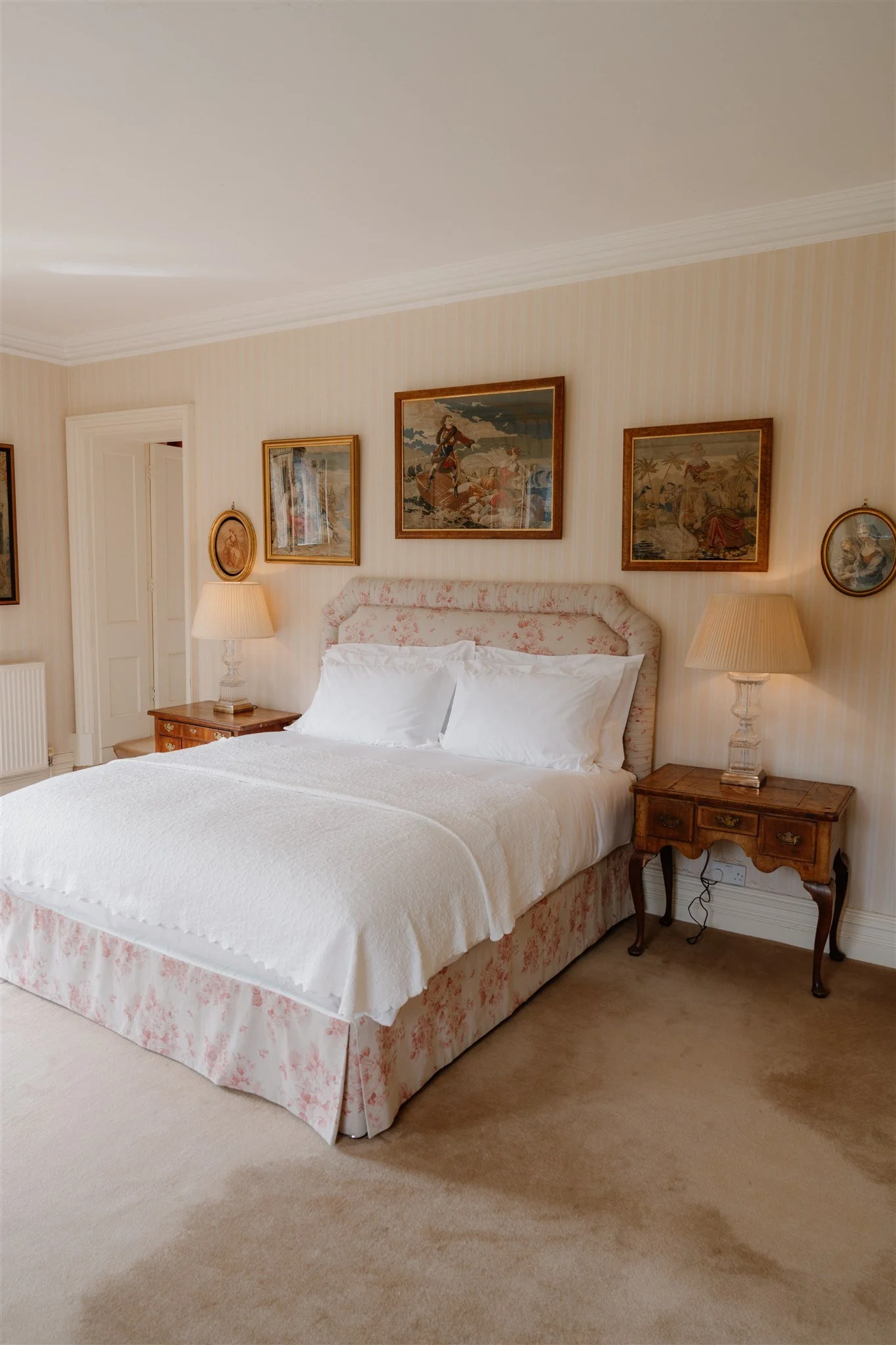 A neatly made white bed with a pink floral bedskirt sits in a traditional bedroom. Two nightstands with lamps are on either side of the bed. Framed artwork hangs on the striped wallpapered wall behind the bed. The room has beige carpeting and a white door to the left.