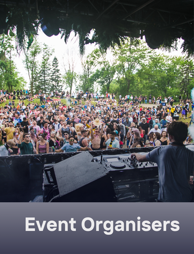 View from a stage showing a DJ DJing at an outdoor music festival with a large crowd of attendees, surrounded by trees and greenery.
