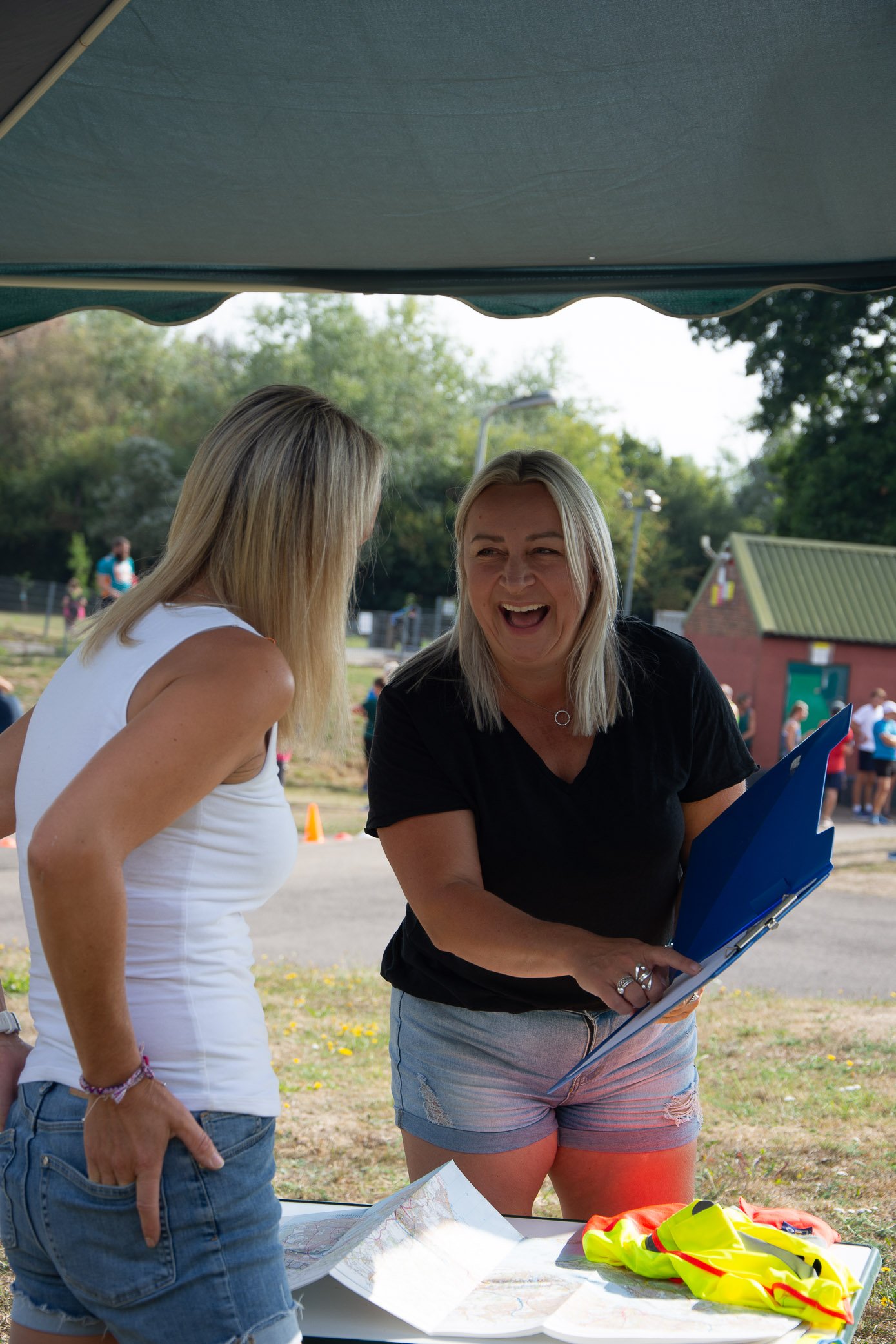Two women are standing outdoors, smiling and talking, with one holding blue folders. They are under a canopy with a park, trees, and people in the background.