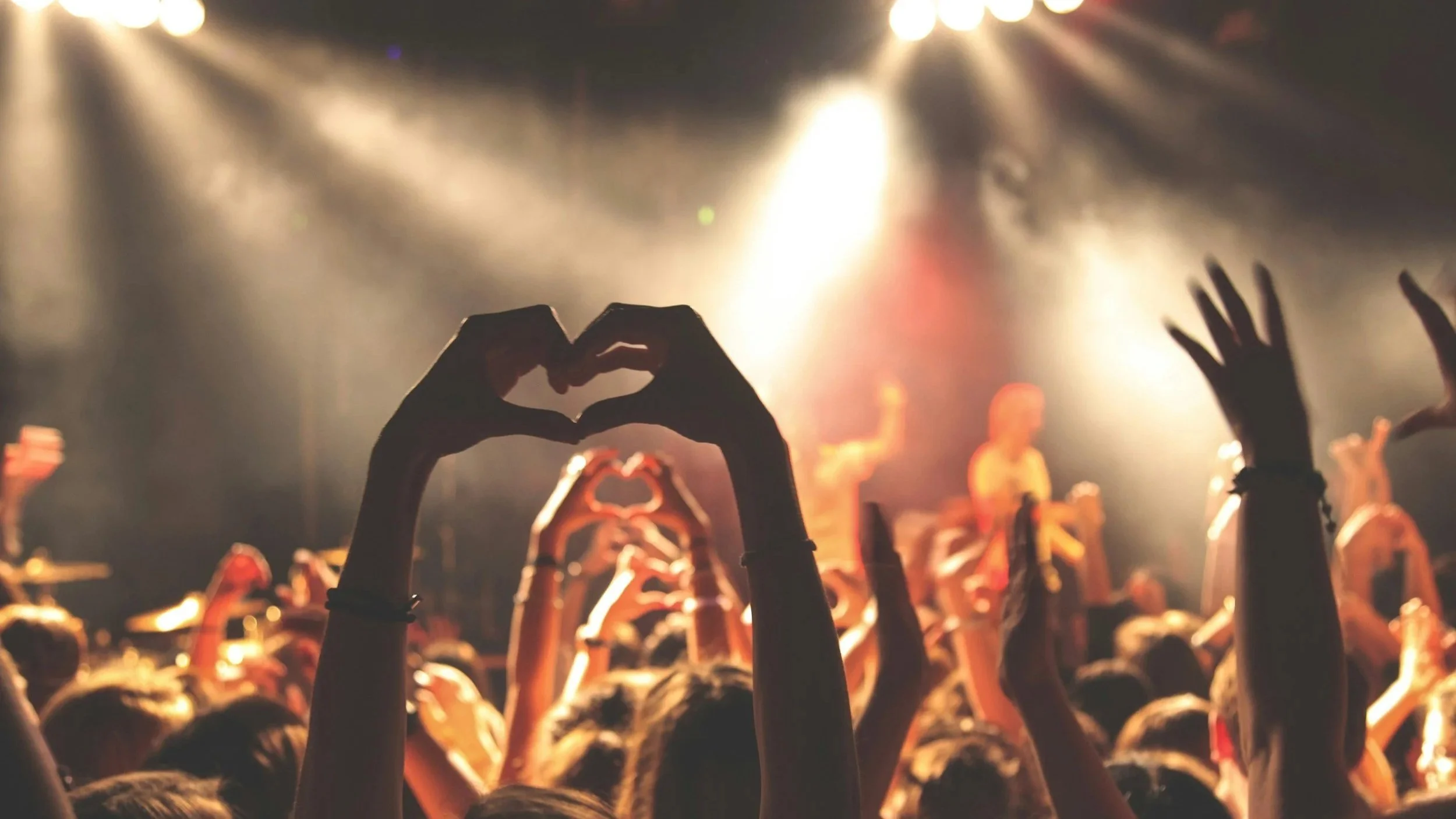 People at a concert showing hands, some making heart shapes, with stage lights in the background.
