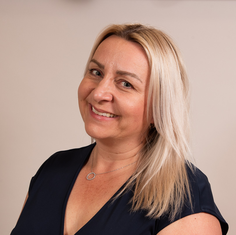 A woman with blonde hair smiling and looking at the camera, wearing a black top and a silver necklace, against a neutral background.