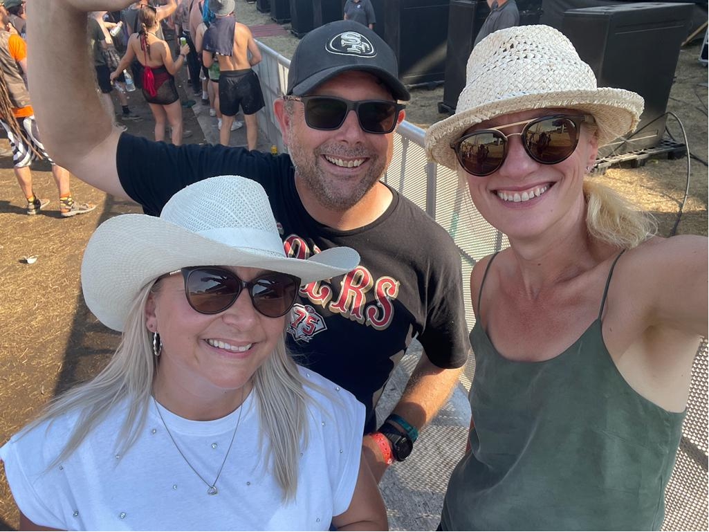 Three smiling people wearing sunglasses and sun hats taking a selfie at an outdoor event with people in the background.