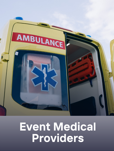 Back of a yellow ambulance with an open door, showing medical equipment inside, and a sign that says 'AMBULANCE' on the rear. The sky is partly cloudy.