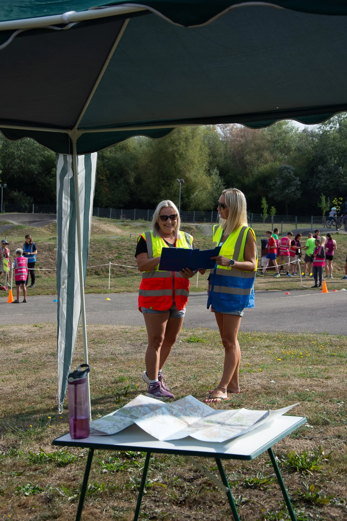 Two women wearing yellow safety vests and shorts looking at a clipboard, standing under a tent outdoors with children in the background.