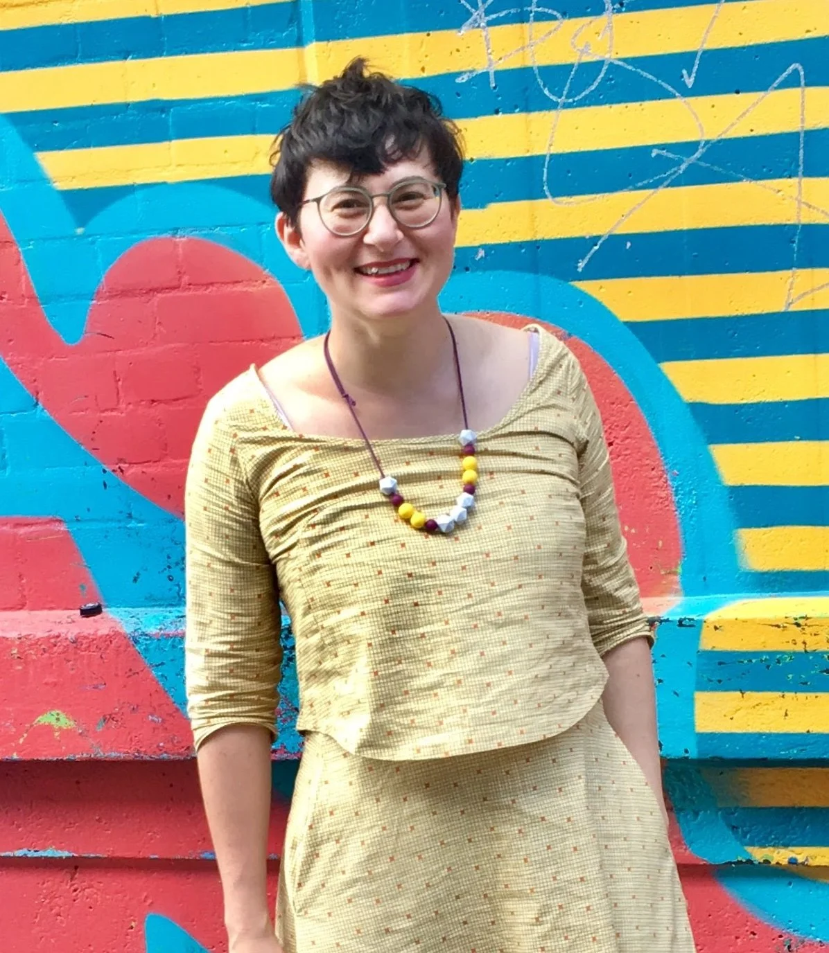 A woman with short dark hair, glasses, and a yellow dress standing in front of a colorful graffiti wall with blue, yellow, red, and white designs.
