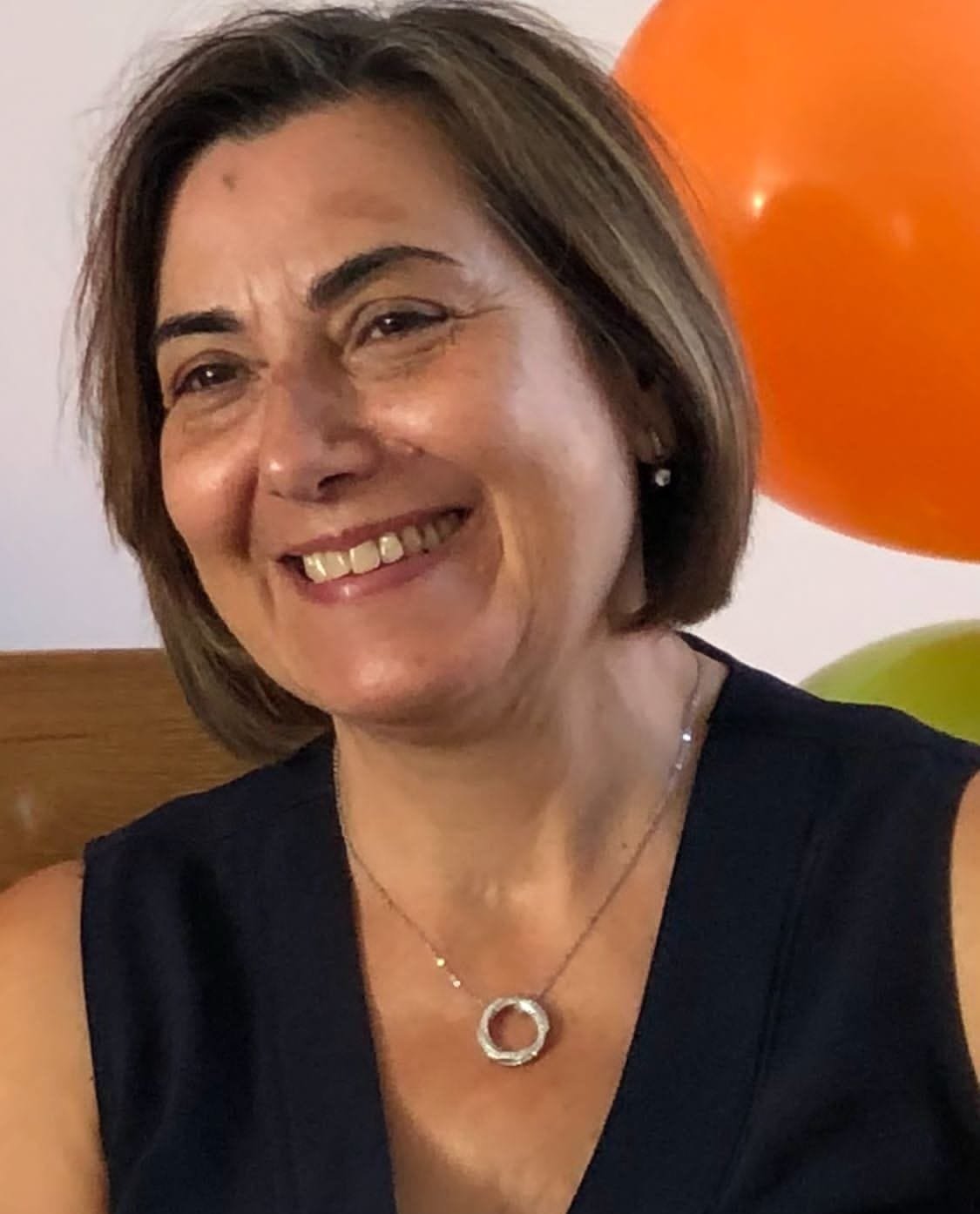 A smiling woman with short brown hair, wearing a black sleeveless top, a necklace, and earrings, standing in front of orange and green balloons on a wall.