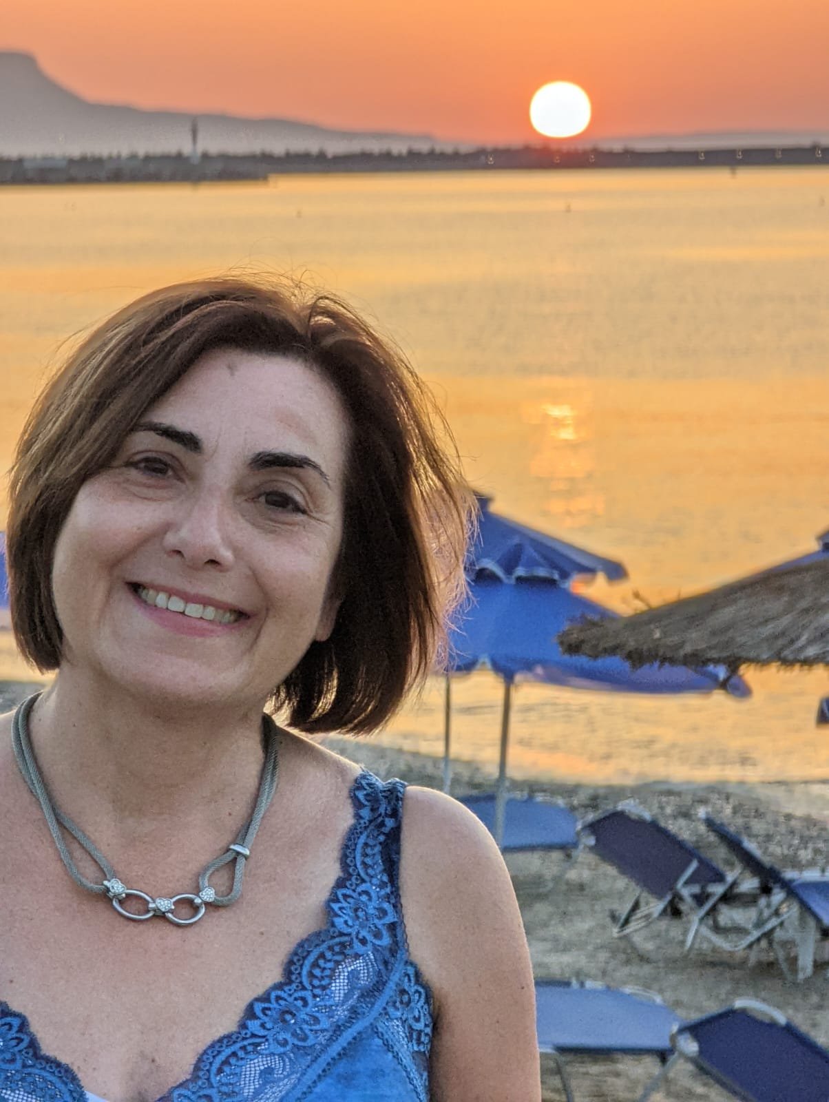 A woman with short brown hair smiling at sunset on the beach, with blue umbrellas and lounge chairs nearby.