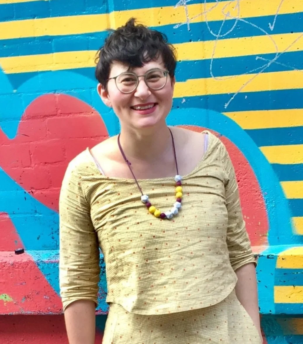 Smiling woman with short dark hair, glasses, wearing a yellow patterned top and necklace, standing in front of a colorful mural with red, blue, and yellow stripes.