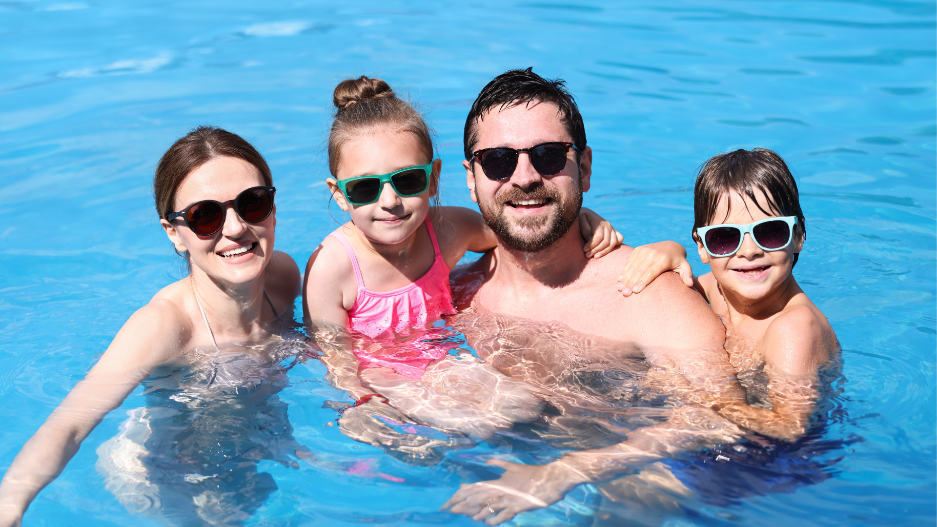 Family of four enjoying a swim in a pool, all wearing sunglasses and smiling