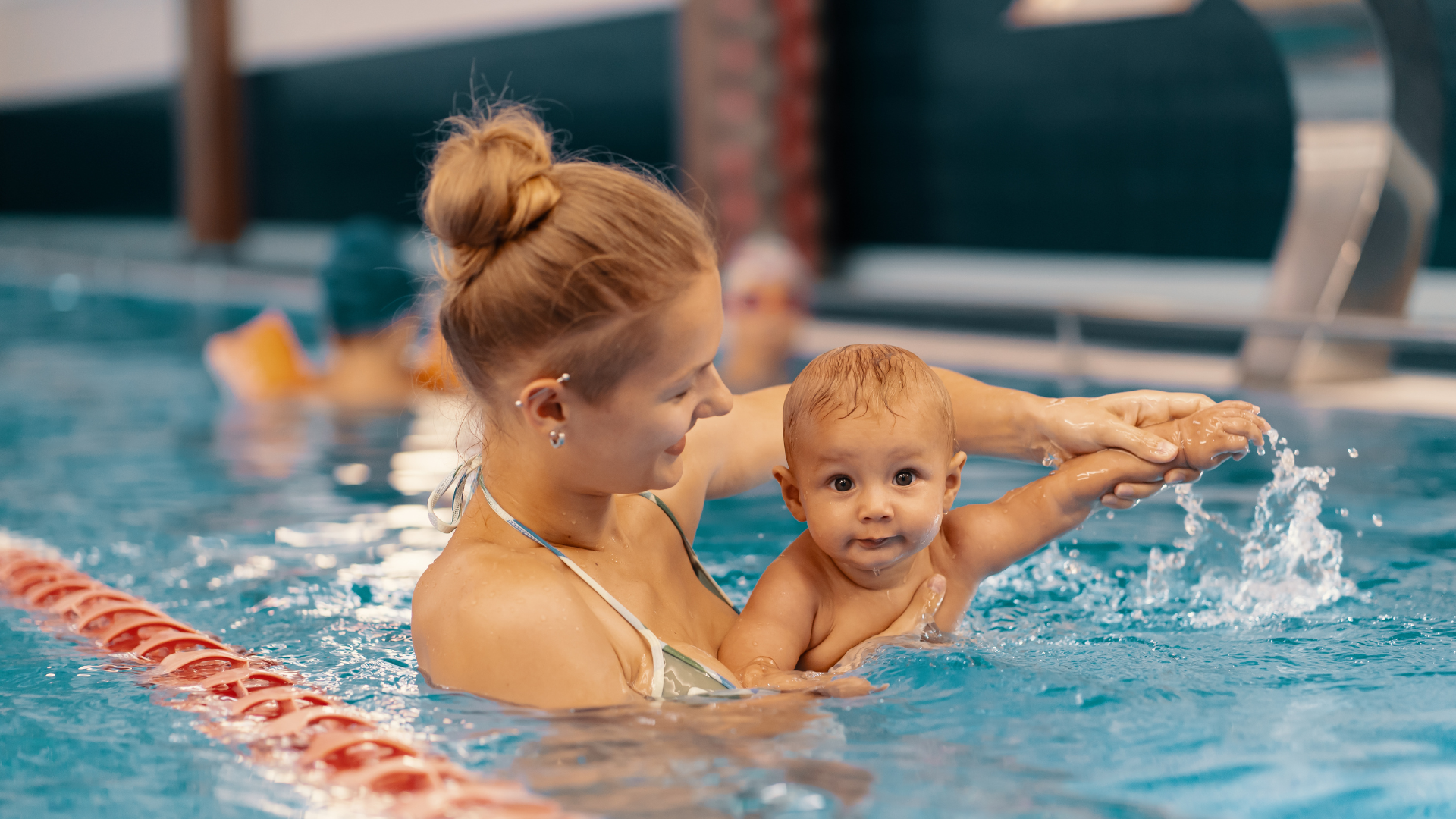 A woman holding a baby in a swimming pool as they practice swimming, with water splashing around them.