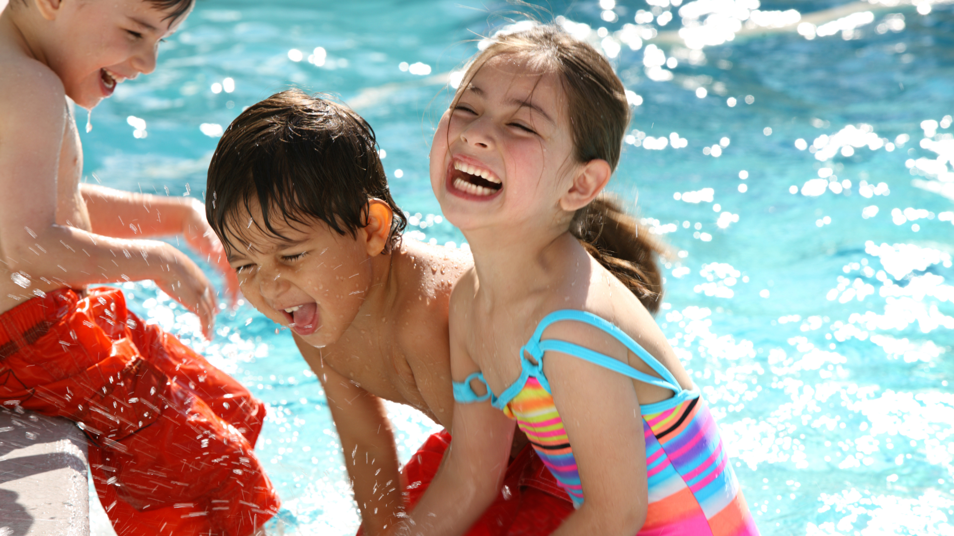 Three children playing and laughing in a swimming pool, splashing water on each other.