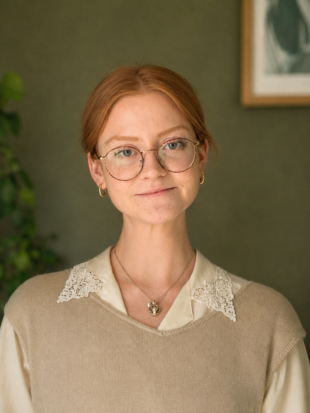 A woman with red hair and glasses wearing a beige sweater with lace details and jewelry, smiling softly.