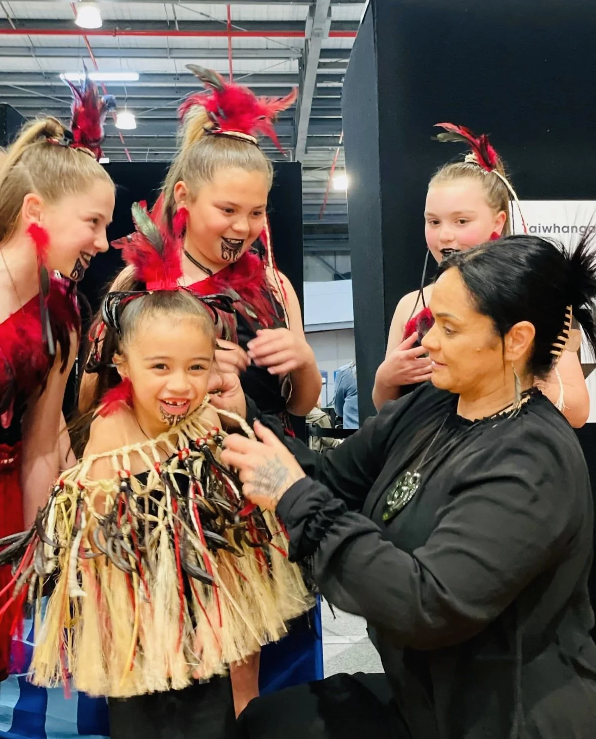 Rangatahi in kākahu backstage at Otago Polyfest while mother adjusts muka cloak before performance.
