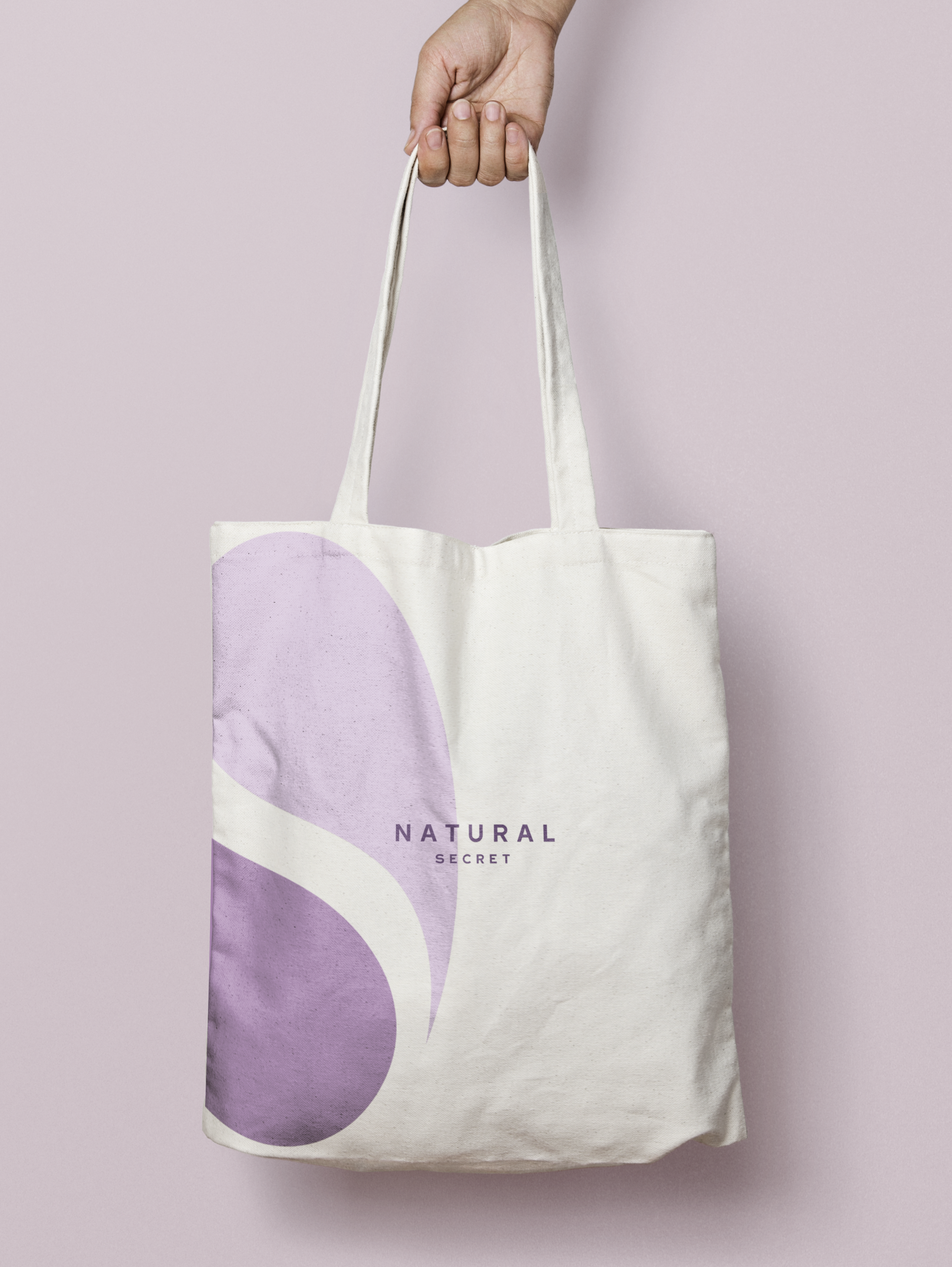 A white tote bag with purple abstract shapes and the words 'NATURAL SECRET' printed on it, hung by a hand against a light pink background.