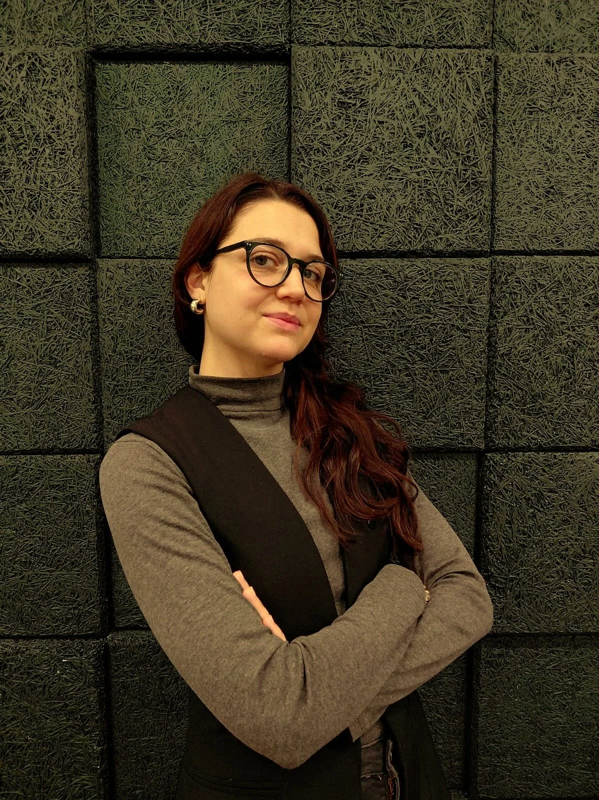 A young woman with long, wavy reddish-brown hair, wearing glasses, a grey turtleneck, and a black sleeveless vest, standing with arms crossed in front of a textured black wall.
