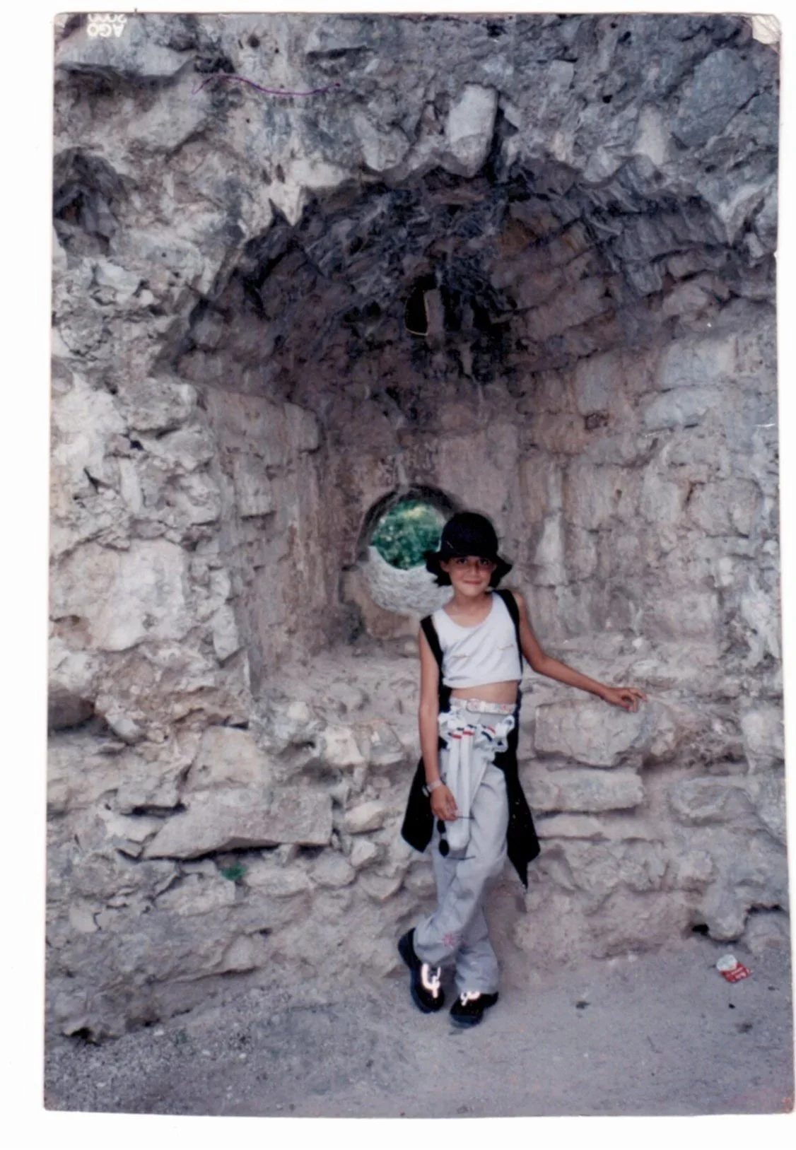 A young girl standing in front of an ancient stone tunnel, wearing a black hat, glasses, a sleeveless top, and light-colored pants, with a smile and a backpack.